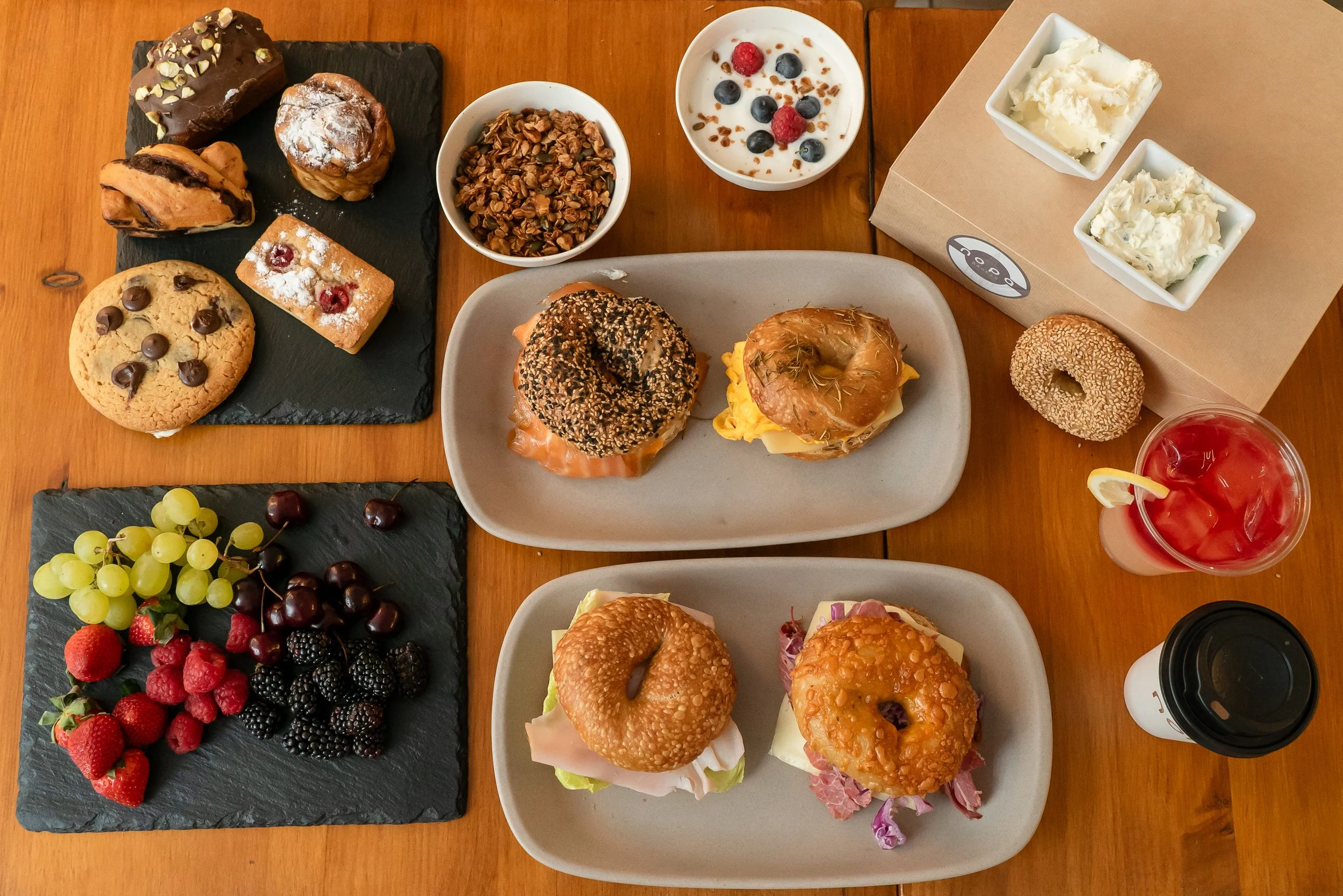 A table with various breakfast items including two plates of sandwiches with bagels, a cup of yogurt with berries, a bowl of granola, a bunch of mixed berries, assorted cookies, muffins, a bagel, a donut, a sweet pastry, a cup of coffee, a glass of pink drink with ice and lemon, and small bowls of butter.