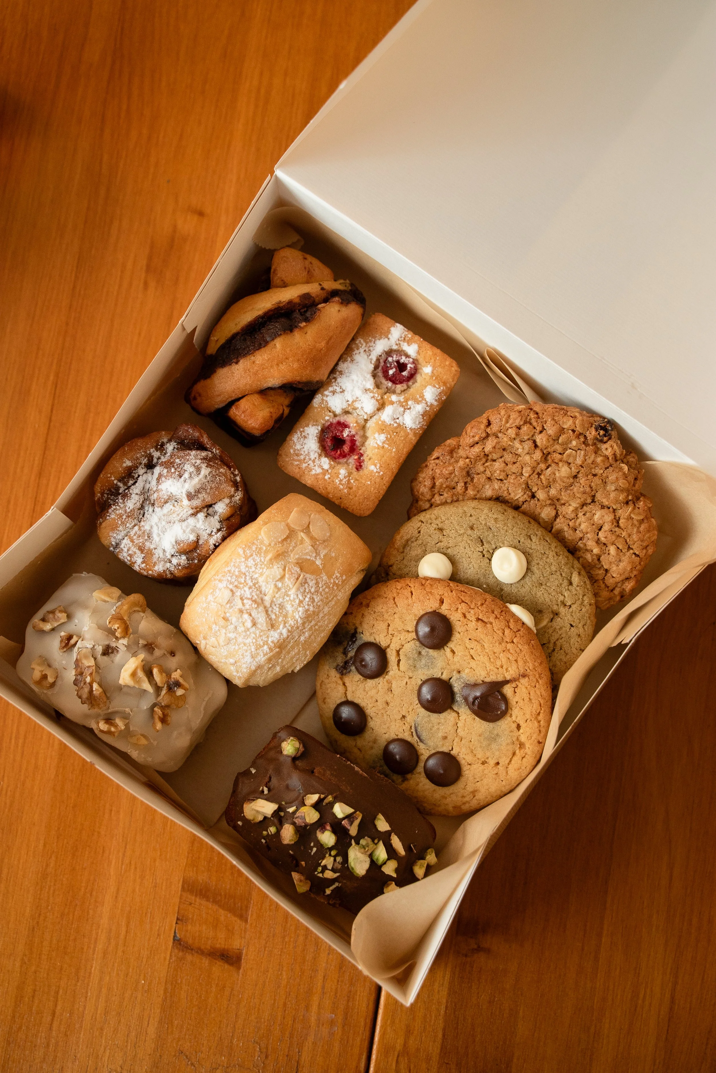 A box of assorted cookies, including chocolate chip, oatmeal raisin, sugar cookies with fruit, and other varieties with toppings, arranged in a white box on a wooden surface.