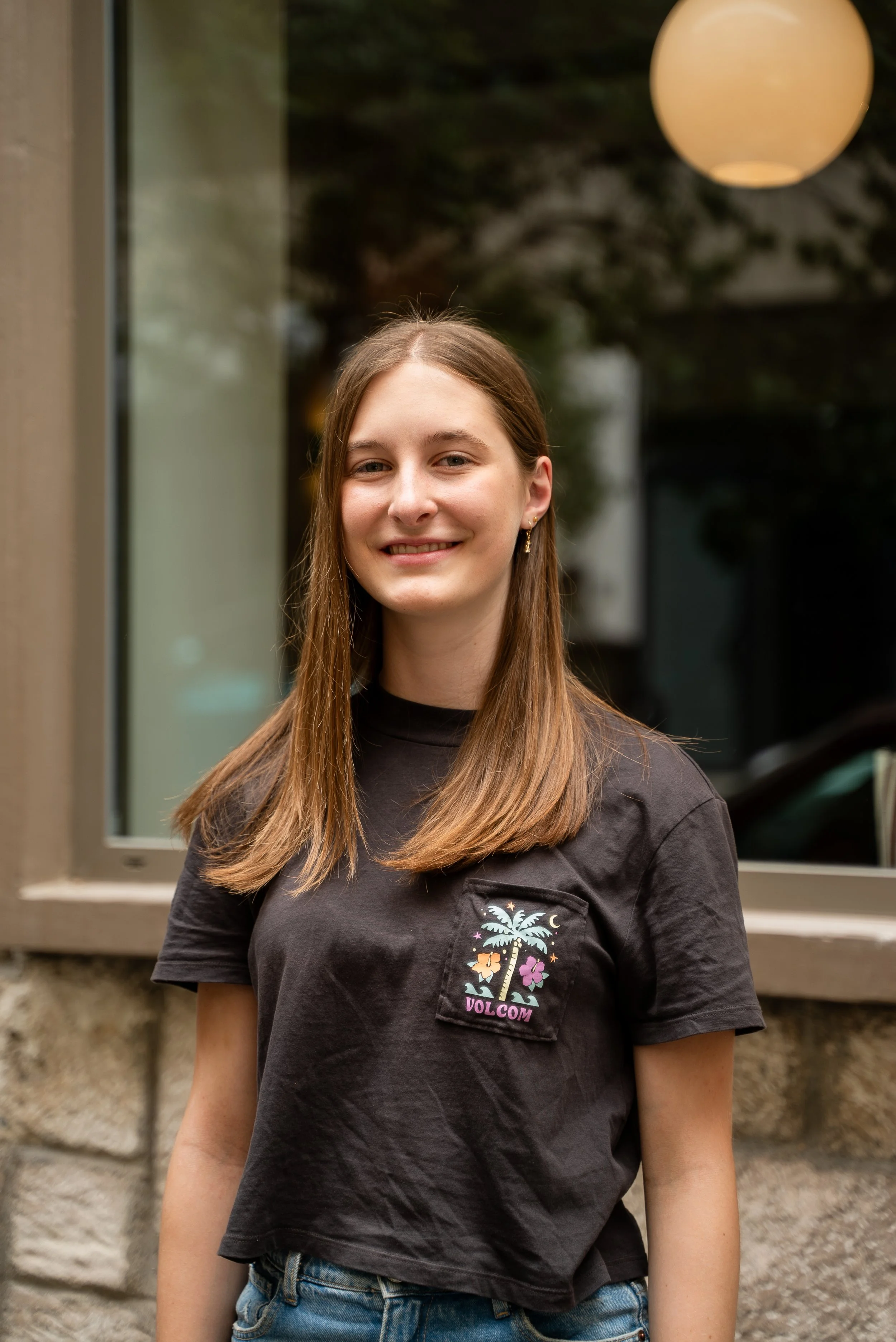 Young woman with long brown hair wearing a black t-shirt with a colorful palm tree logo, standing outside near a window and smiling.