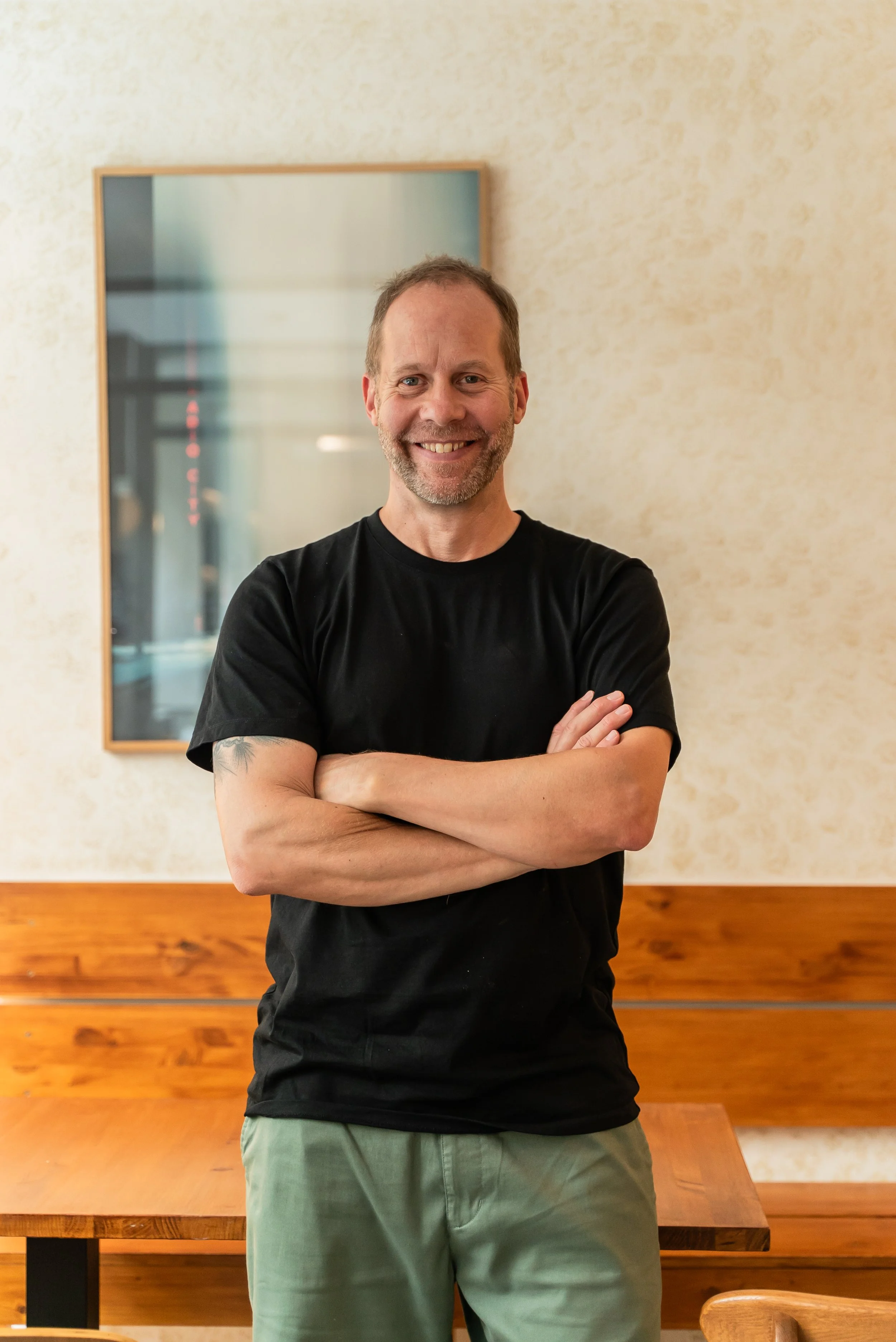 A smiling man with a beard in a black t-shirt and light green pants standing with arms crossed in a room with beige walls and wooden furniture.
