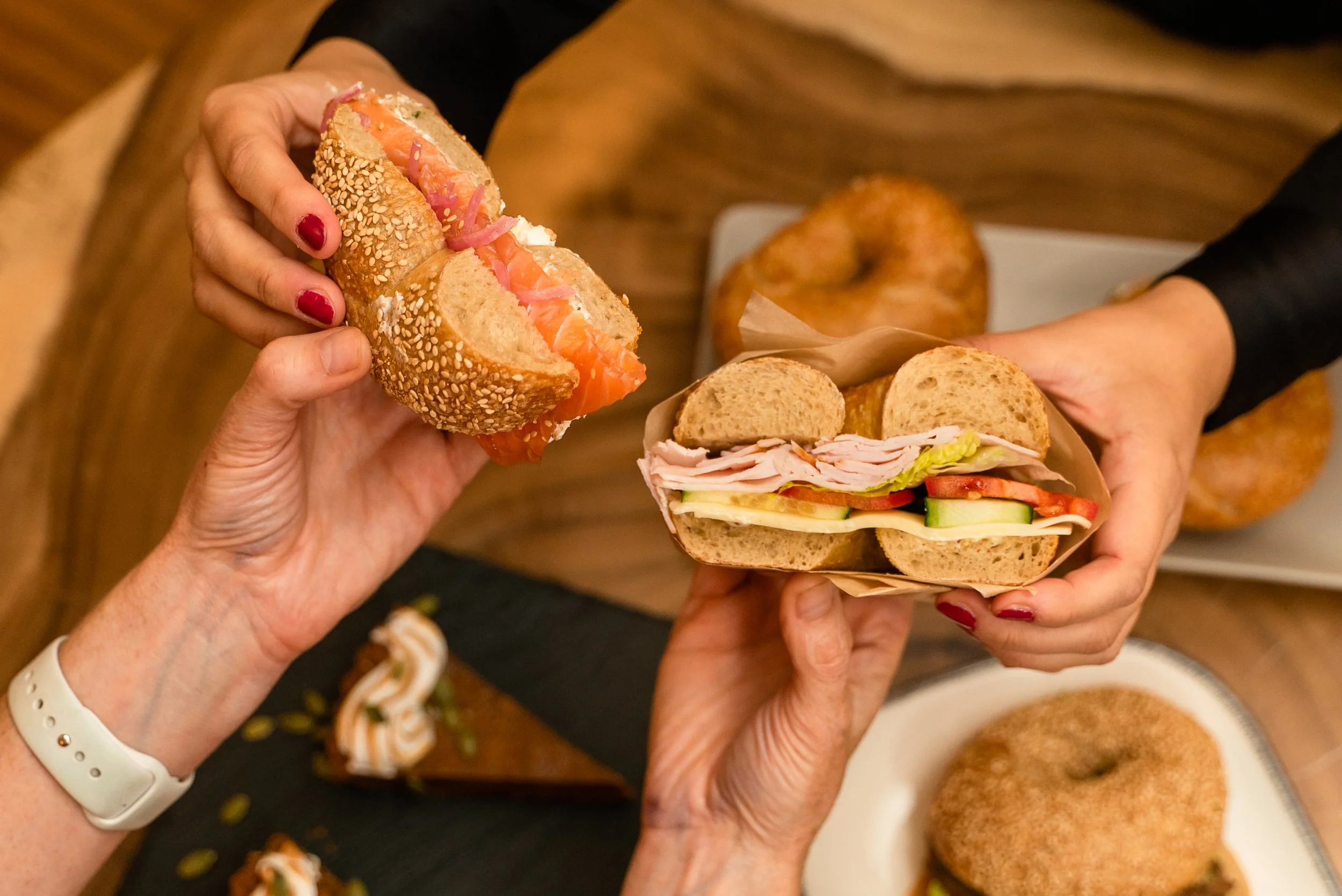 Two people holding sandwiches with various breads, vegetables, and meats, with bagels and desserts on a wooden table in the background.