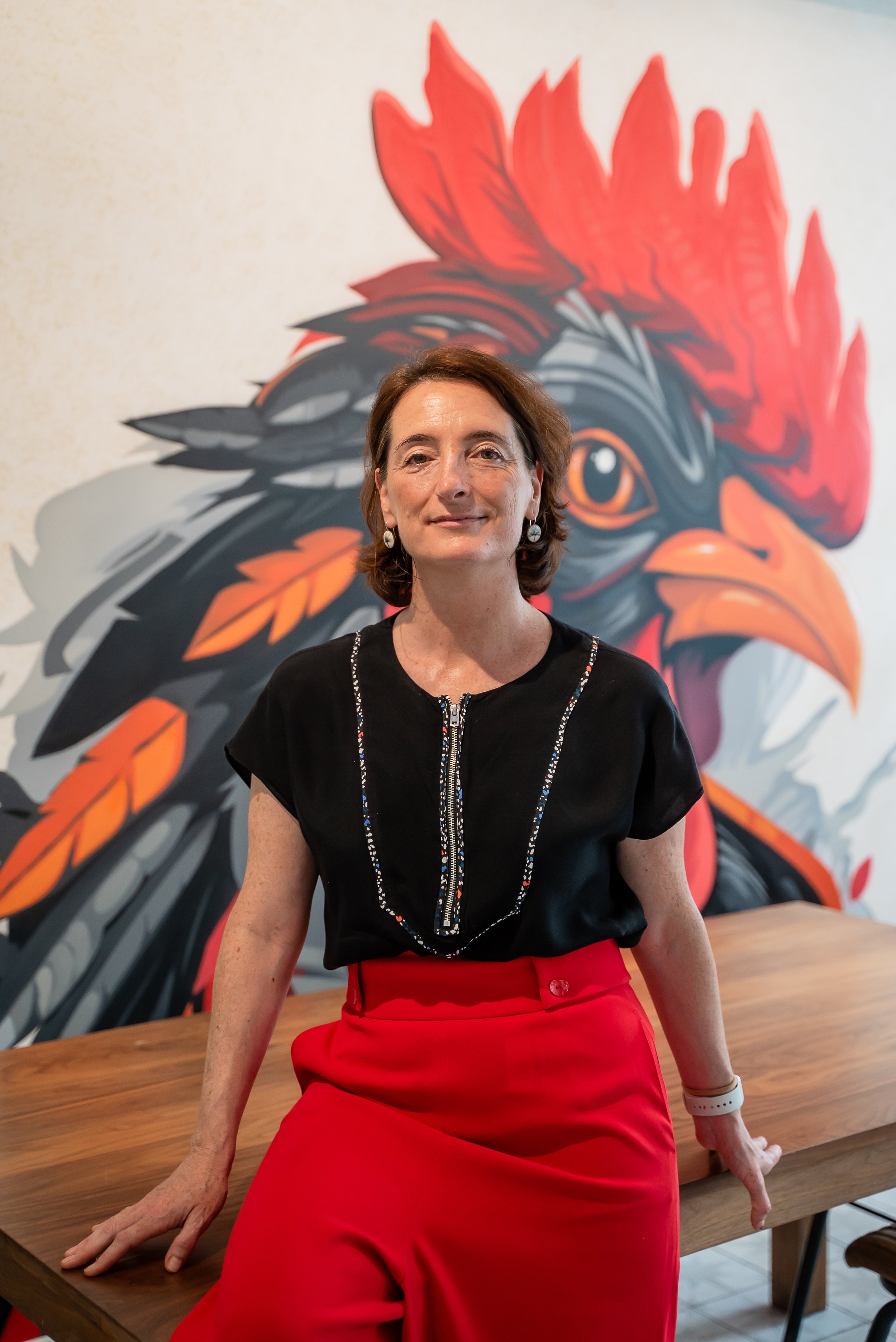 A woman with short brown hair wearing a black top and a red skirt, sitting at a wooden table, with a large colorful rooster mural in the background.