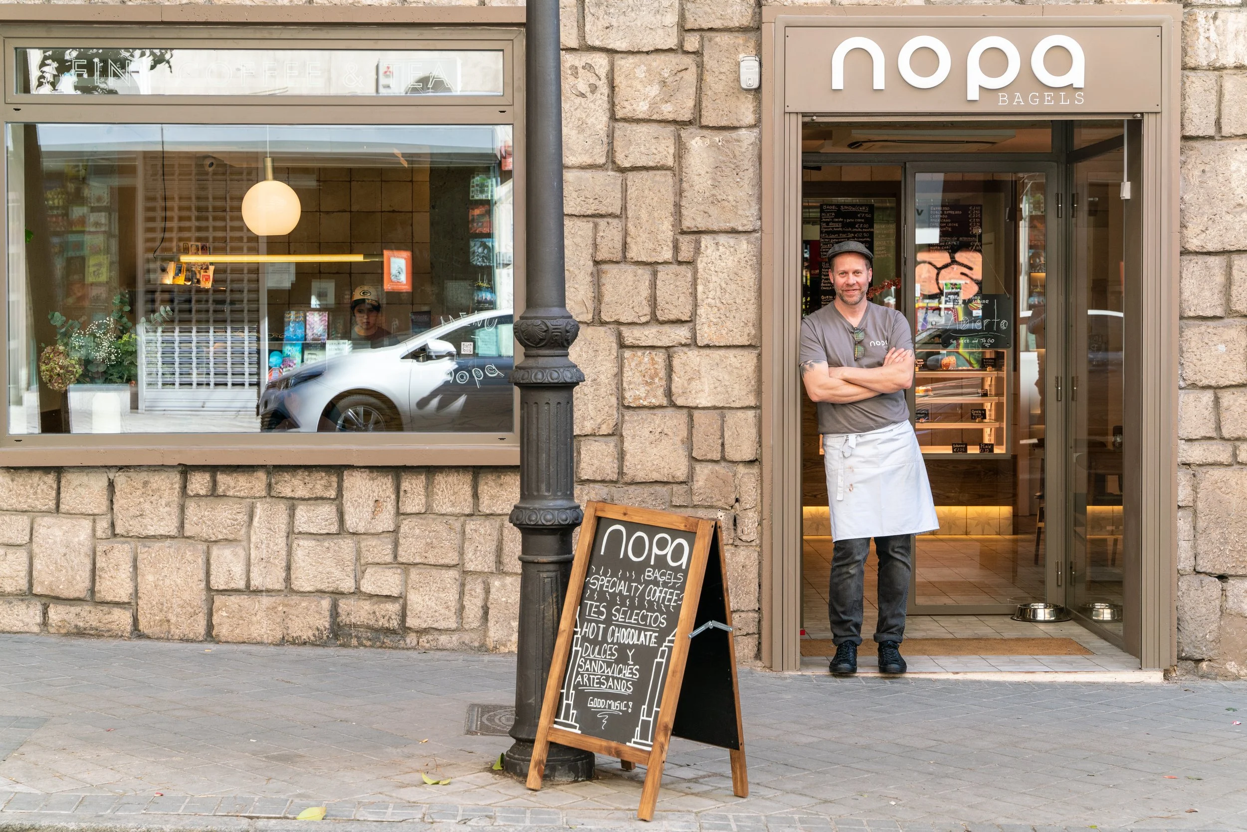 A man standing in the entrance of a bakery named 'NoPa Bagels,' with a chalkboard menu outside listing offerings like coffee, teas, chocolate, sweets, and sandwiches. The bakery has a stone exterior and large window, with a reflection of a car passing by.