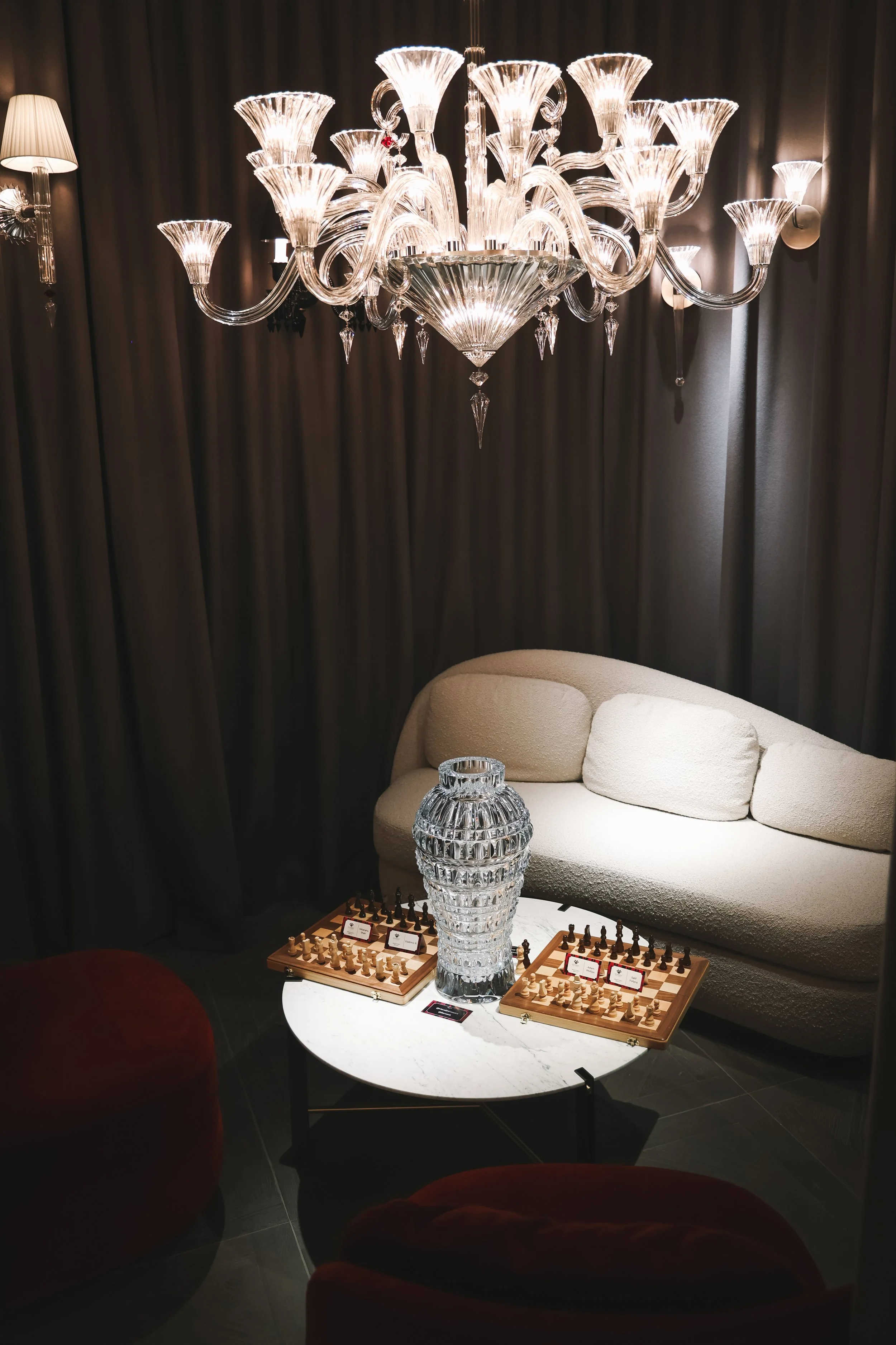 A chandelier with multiple glass arms and light bulbs hangs above a small round table. The table has a large decorative glass vase and two chessboards with pieces. A beige sofa with cushions is in the background, with dark curtains behind the chandel
