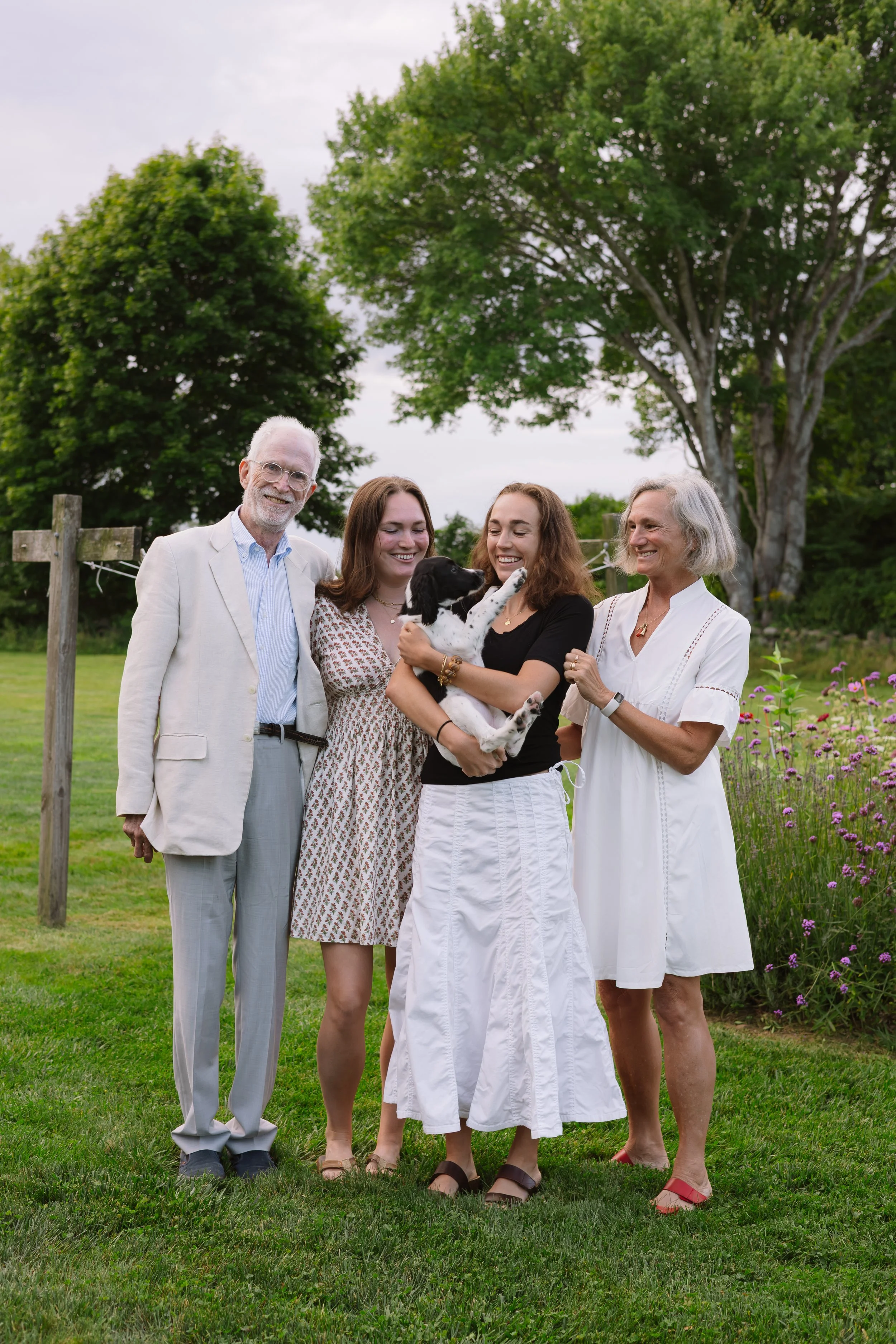 A group of five people standing outdoors on a lawn with trees in the background, smiling and holding a black and white puppy.