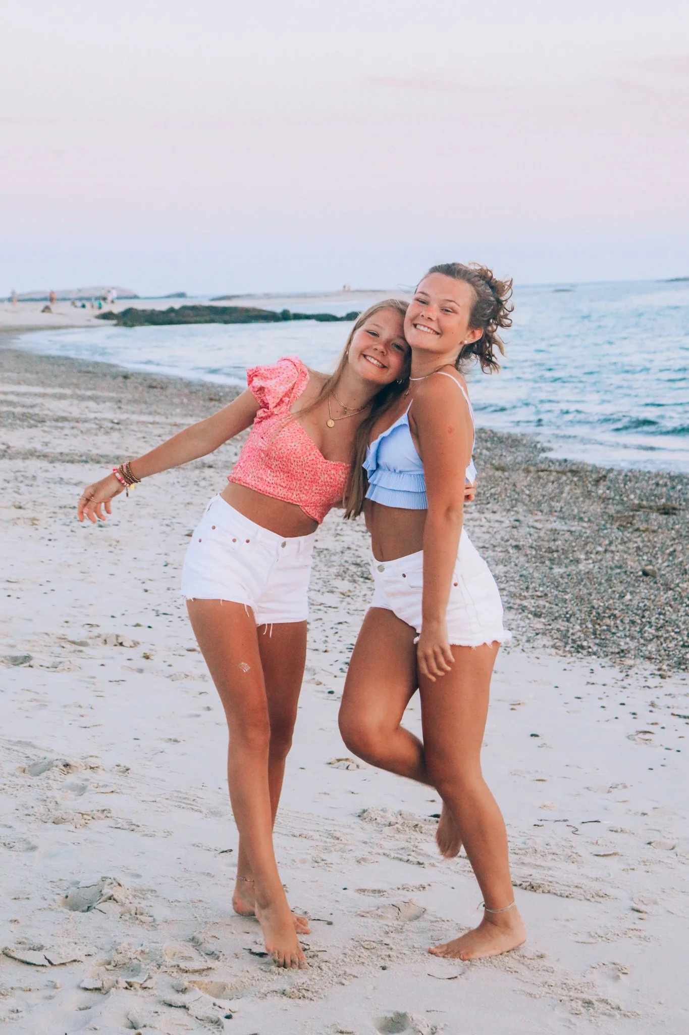 Two young women in summer clothes smiling and hugging on a beach at sunset.