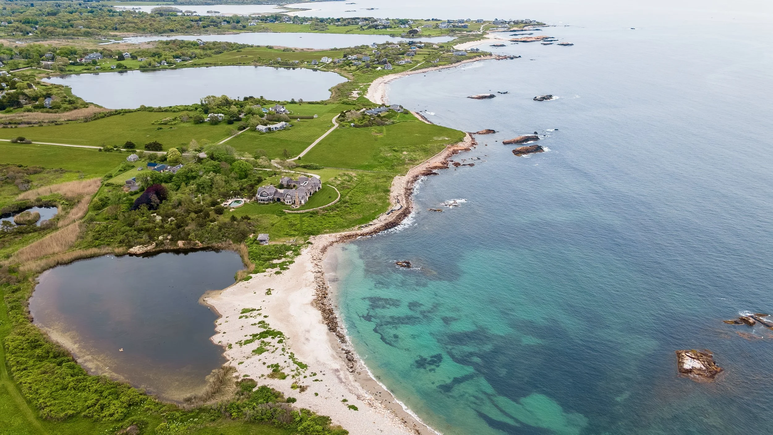 Aerial view of a coastal landscape featuring a mix of homes, lush green fields, ponds, and a rocky shoreline with turquoise water, small waves, and scattered rocks.