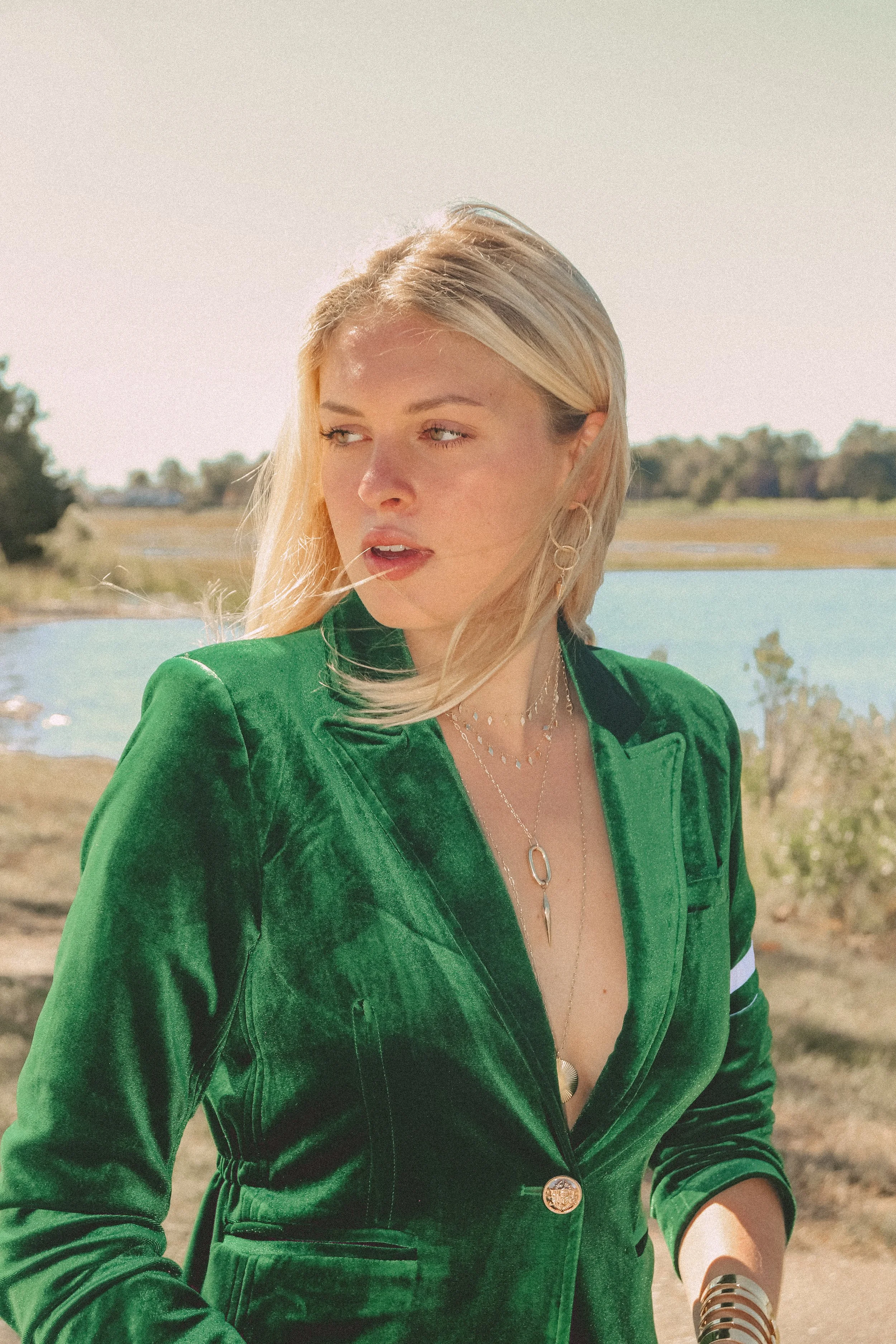 A woman with blonde hair wearing a green velvet blazer, silver jewelry, and a black choker, standing outdoors next to a body of water on a sunny day.