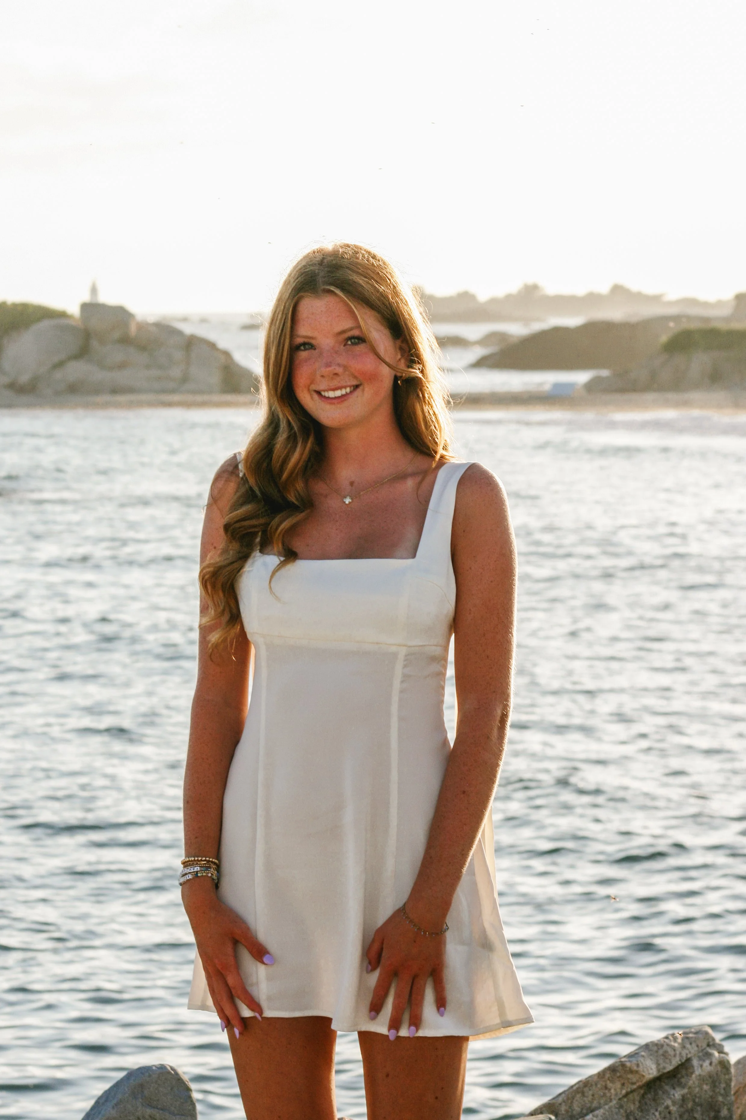 A young woman with long, wavy auburn hair, smiling and wearing a white sleeveless dress, stands on rocks by the water with a rocky shoreline and lighthouse in the background during sunset.