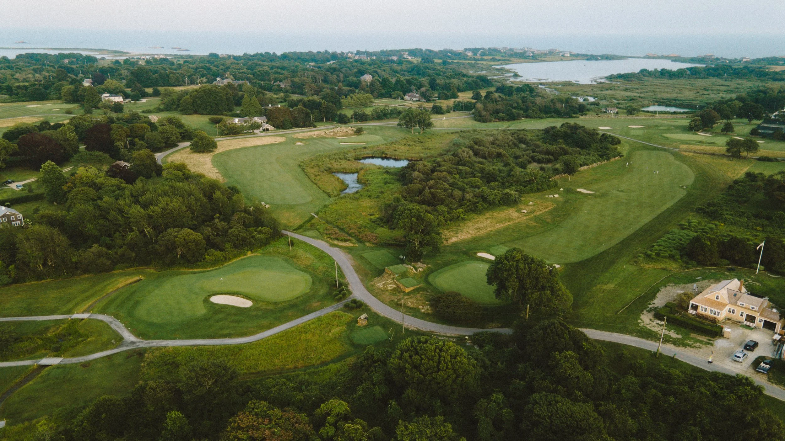 Aerial view of a golf course with several greens, sand traps, water hazards, sand traps, and surrounding trees and residential houses.