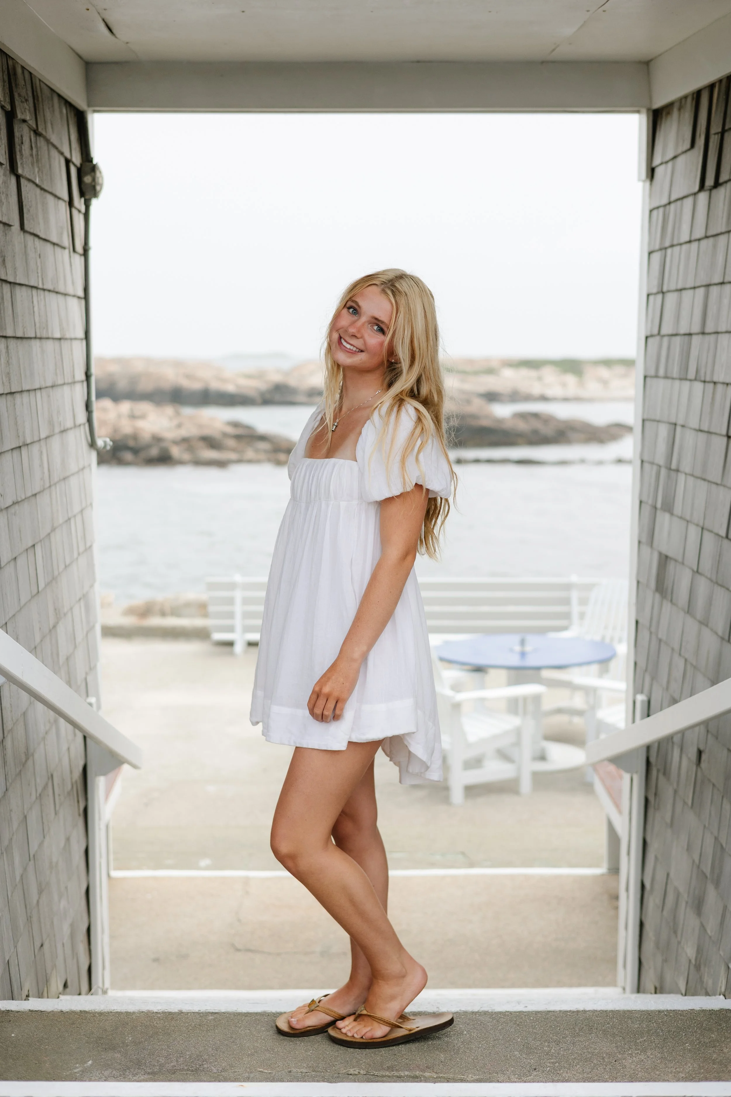 A young woman with long blonde hair smiling, wearing a white dress and flip-flops, standing at the entrance of a beachside pavilion, with rocks and water in the background.