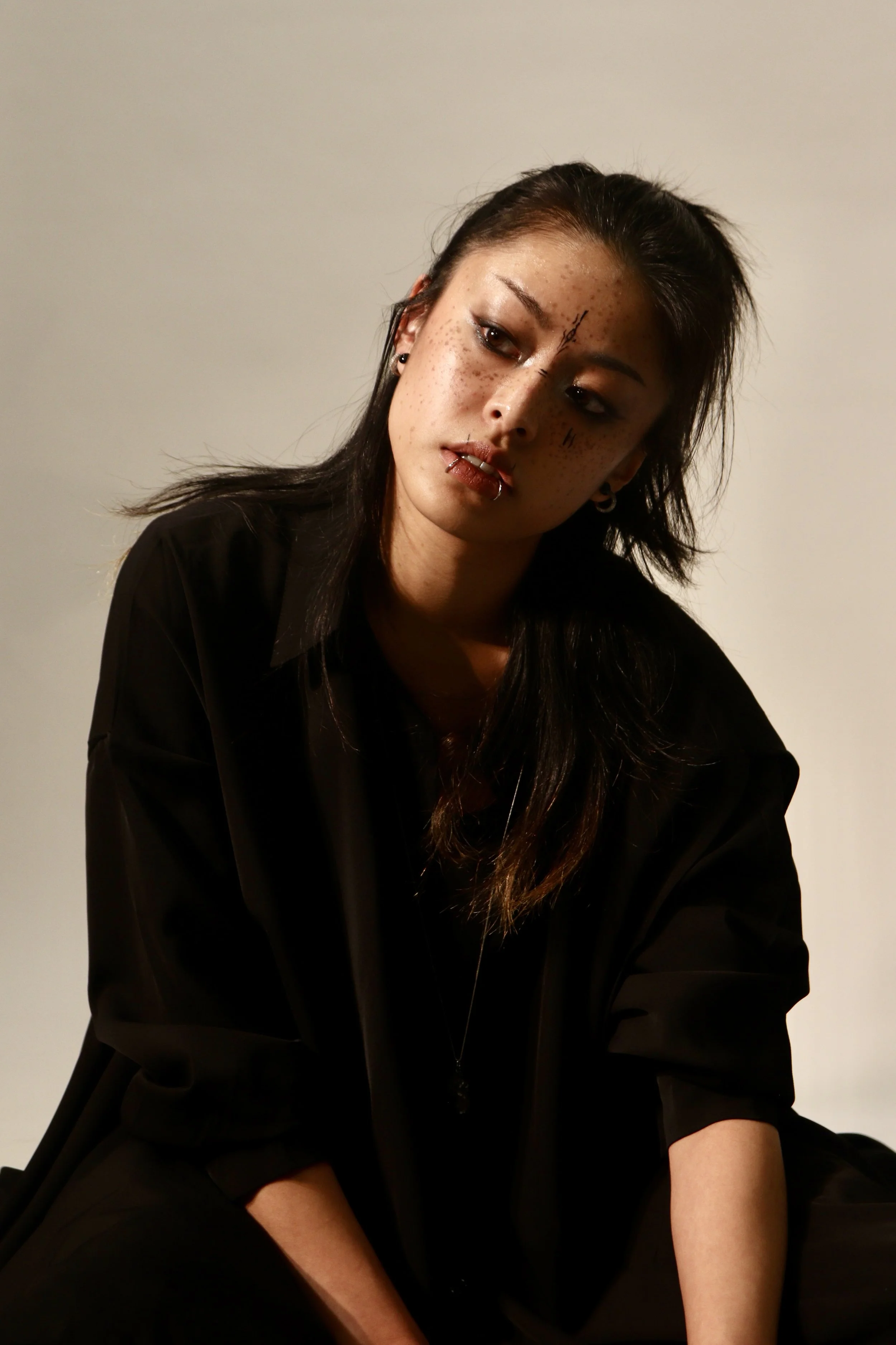 A young woman with dark hair, freckles, and makeup with a gothic style, wearing a black shirt, sitting against a plain background, looking down with a serious expression.