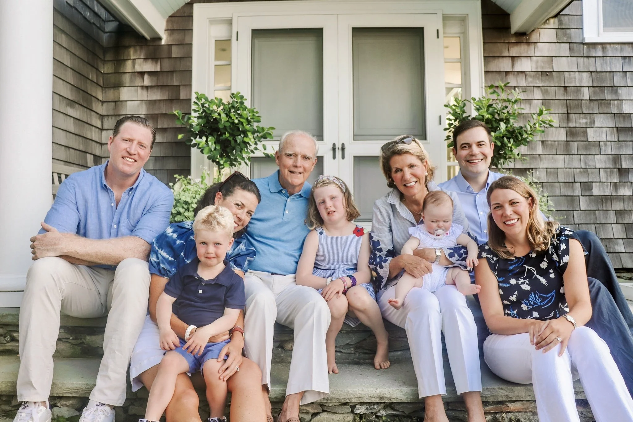 A multigenerational family sitting on the steps of a porch outside a house with a wooden exterior and a white door. They are smiling and posing for the photo.