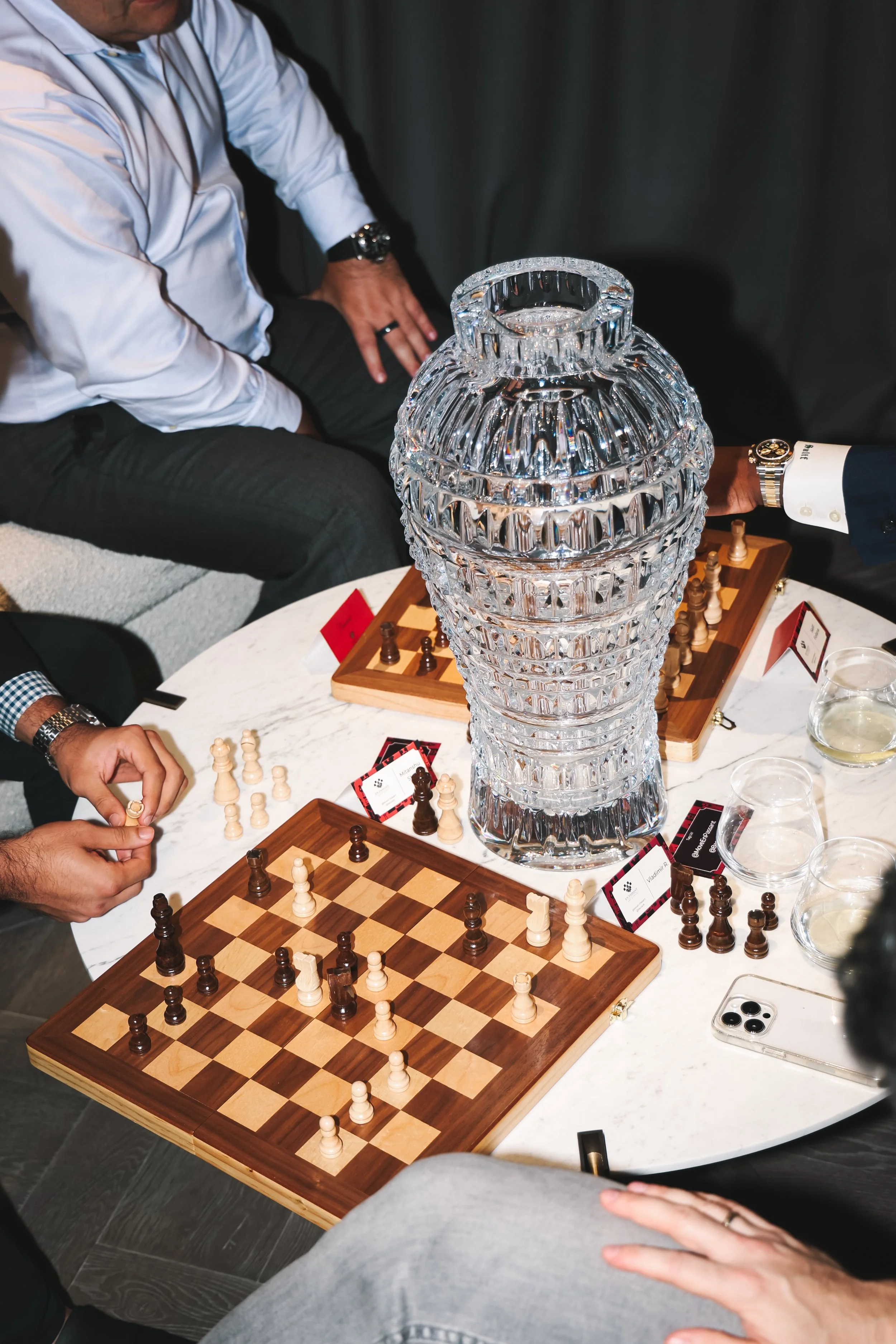 People playing chess at a table with a large glass sculpture and chess set, with cards and drinks nearby.