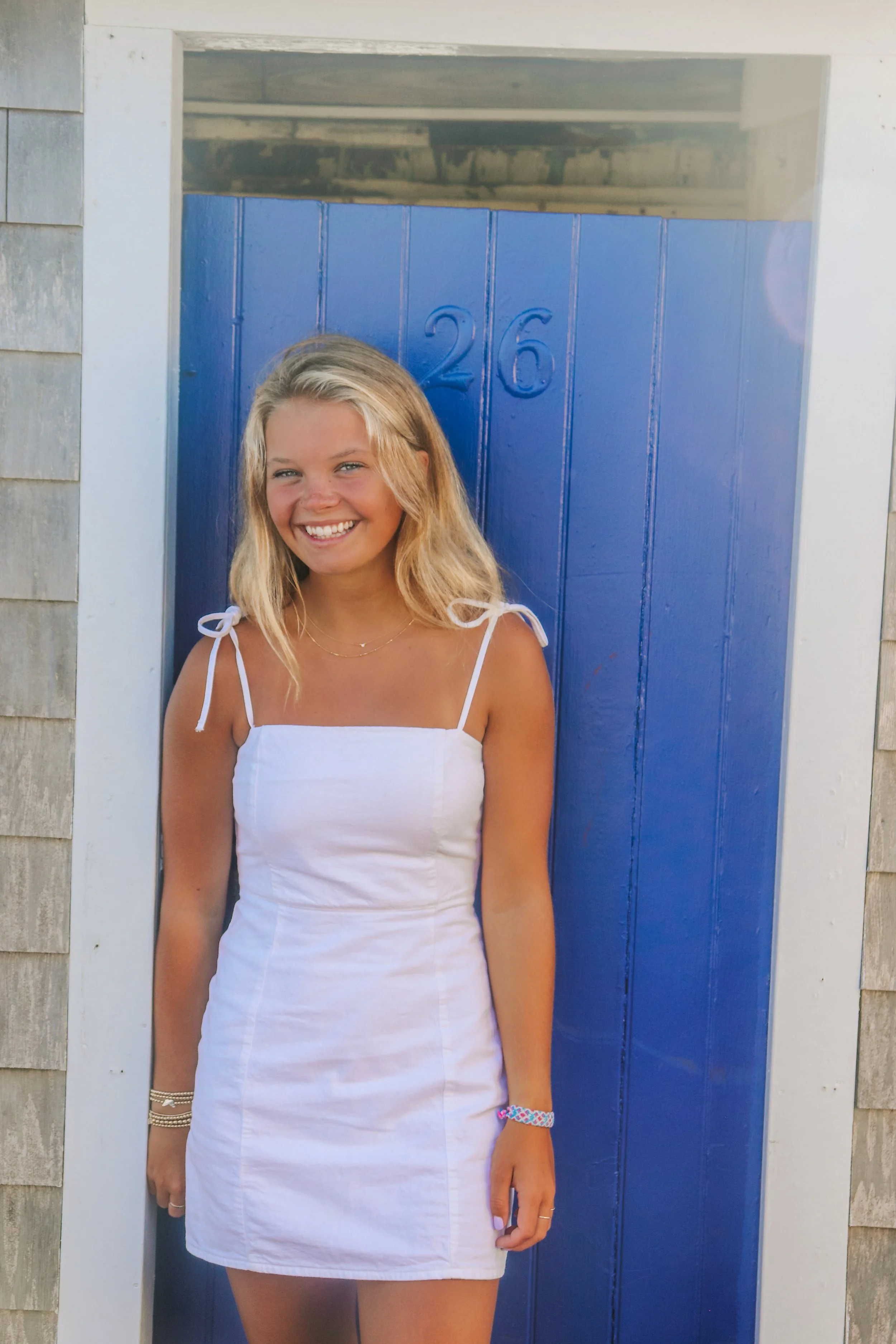 A young woman with blonde hair, wearing a white dress with tied straps, standing in front of a blue door with the number 26, outside a house with wooden shingles.