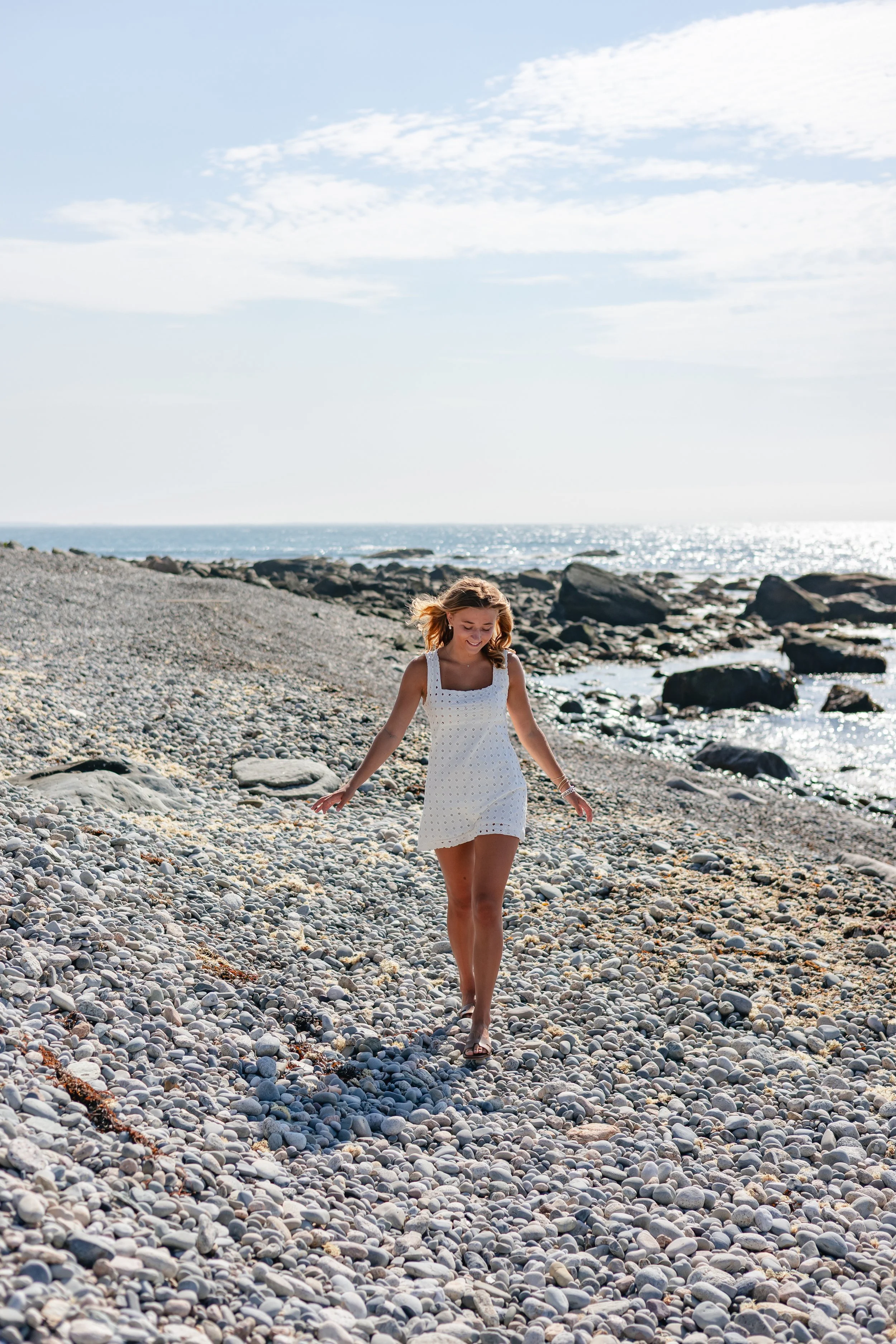 A woman walking along a pebble beach with rocks and the ocean, smiling, under a partly cloudy sky.