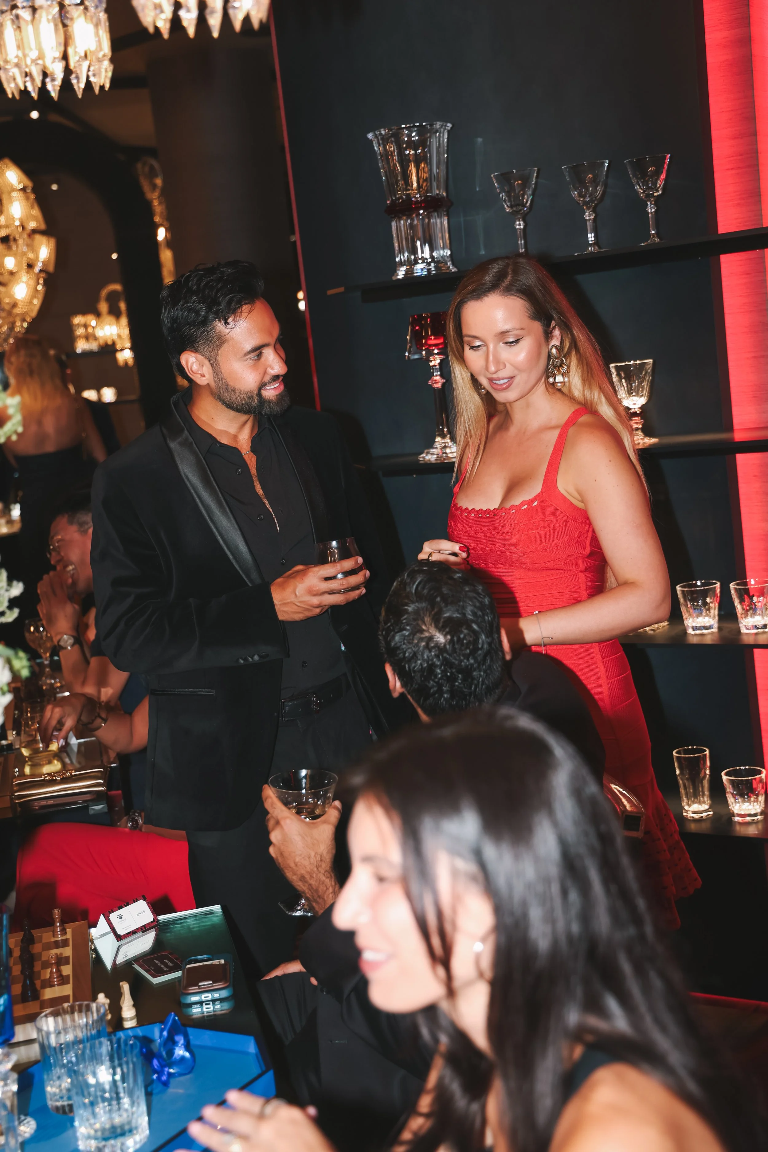 People socializing and playing chess at a formal gathering or event, with one woman in a red dress and a man in a black suit holding a drink in a luxurious setting with chandeliers and decorative glassware.