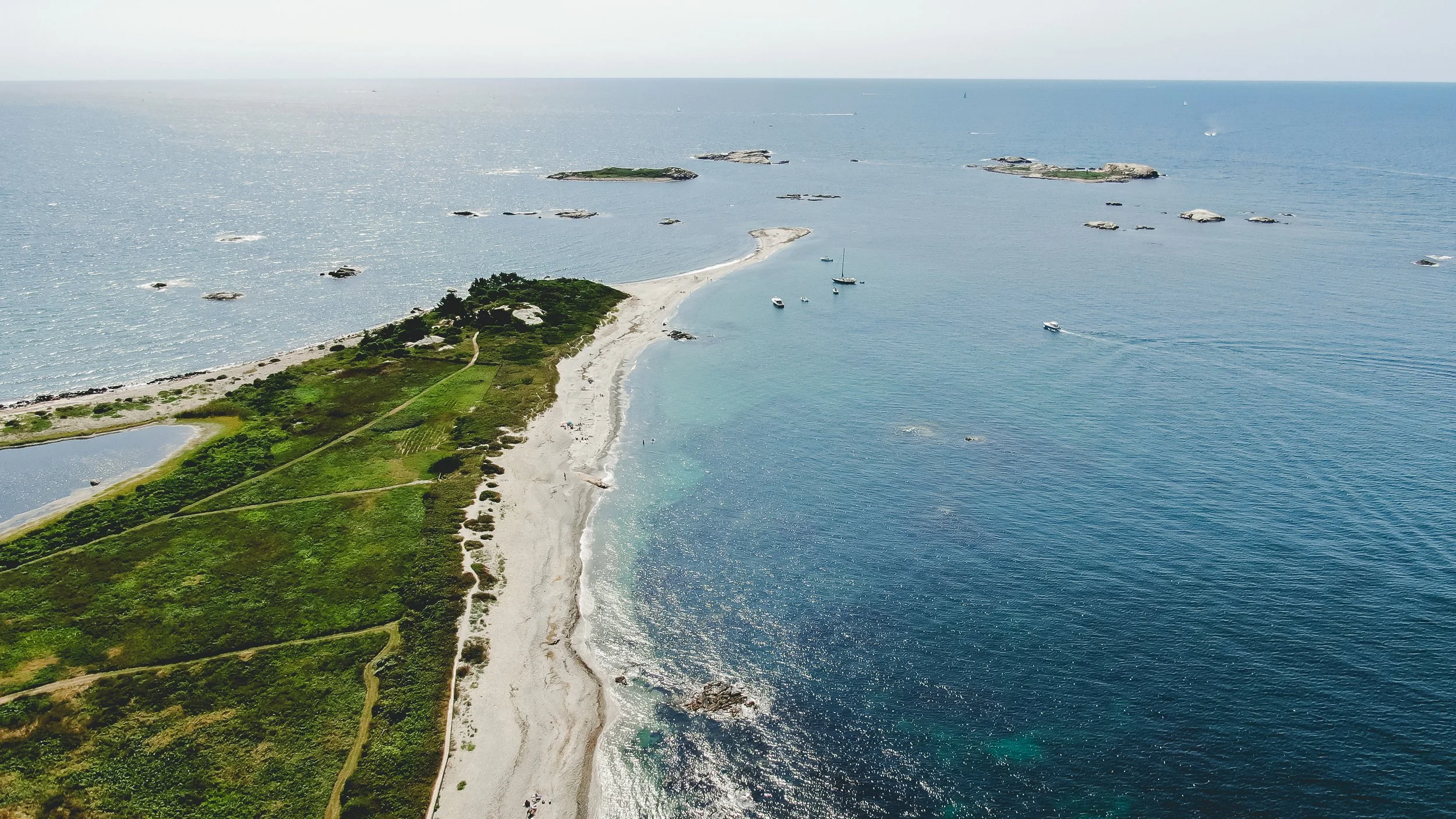 Aerial view of a coastal landscape with a sandy beach, green vegetation, and several boats in the water, with multiple small islands scattered offshore.