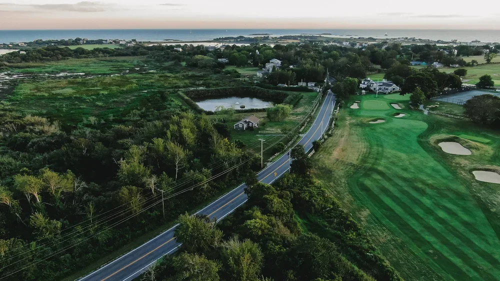 An aerial view of a rural landscape featuring a winding road, a golf course with sand traps, a pond, lush green fields, and scattered houses near the coastline.