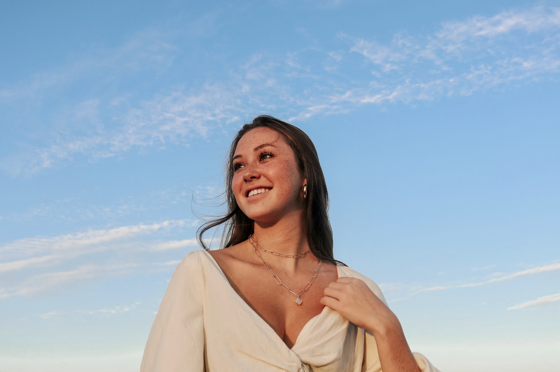 Young woman smiling outdoors with blue sky and clouds in background, wearing jewelry and a white top.