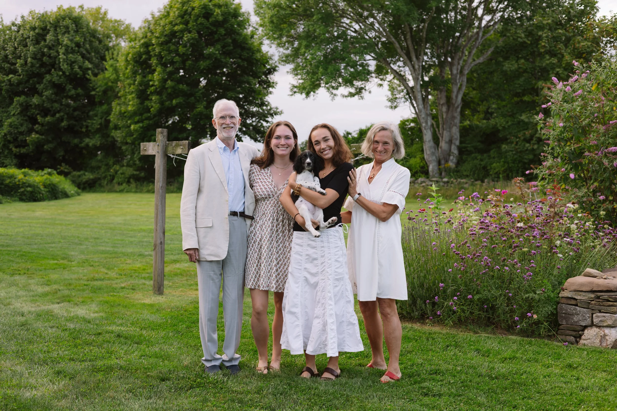 A group of five people and a dog standing outdoors on a grassy area with trees and flowers in the background.