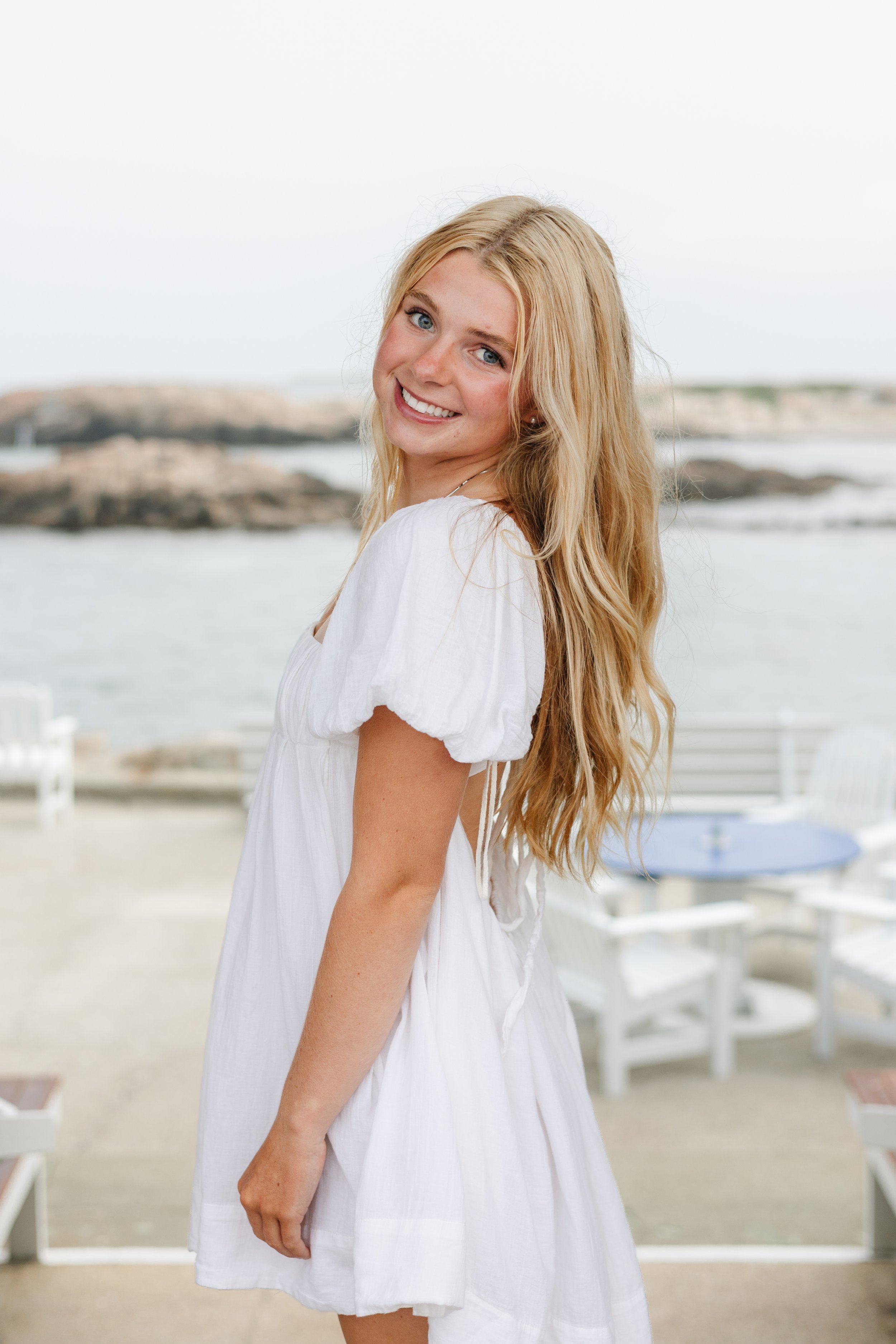 A young woman with long blonde hair and blue eyes smiling at the camera outdoors near the water.