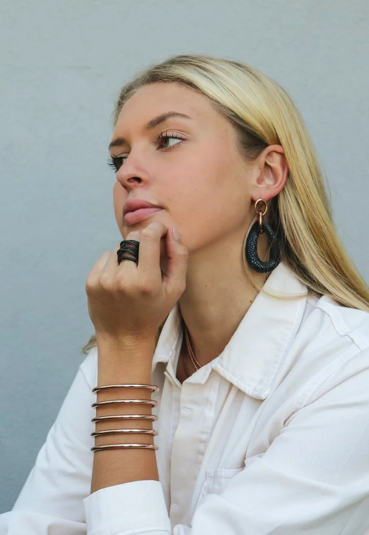 A young woman with blonde hair in a white shirt, wearing large hoop earrings and multiple bracelets, resting her chin on her hand while looking thoughtfully to the side against a plain gray background.