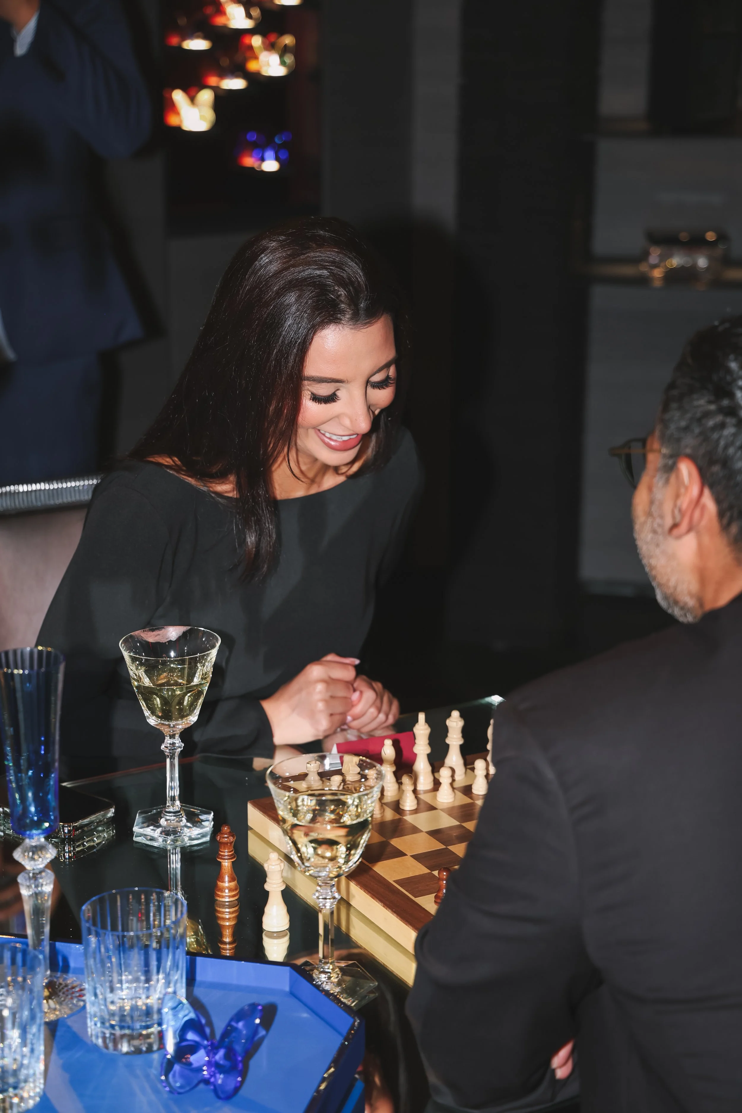 A woman and man playing chess at a table in a dimly lit room. The woman has long dark hair, is smiling and leaning forward with her hands clasped. The man, wearing glasses, is facing away from the camera. The table has chess pieces and glasses of win
