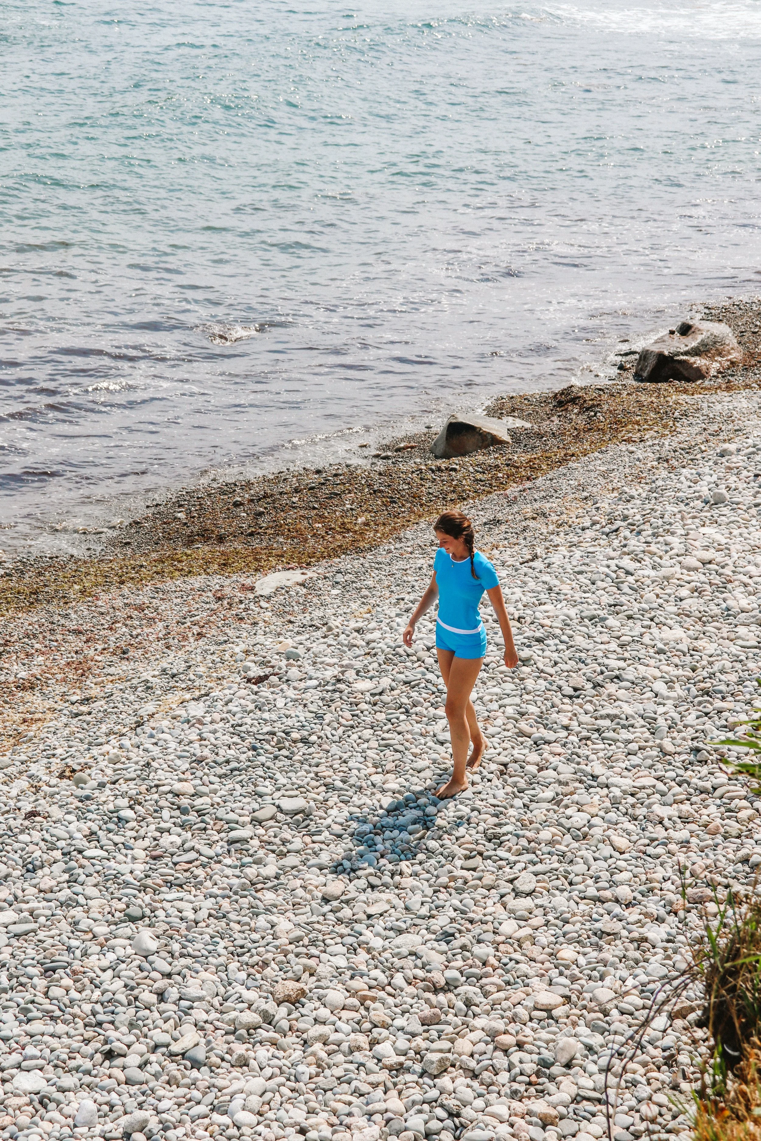 A woman in a blue outfit walking barefoot on a pebbled beach next to the ocean.
