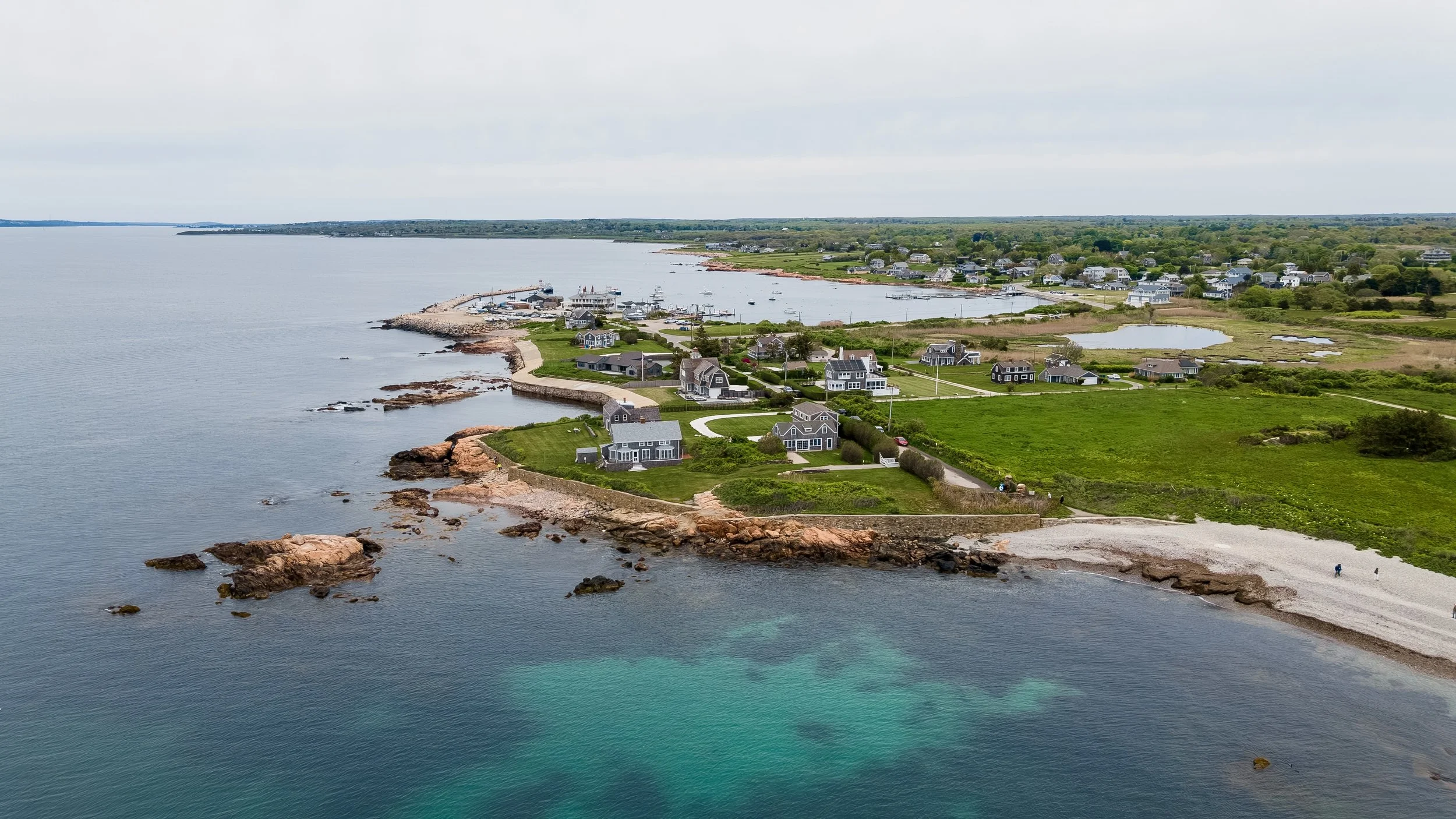 Aerial view of a coastal community with houses near the shoreline, a small harbor with boats, rocky beaches, and green hills under a cloudy sky.