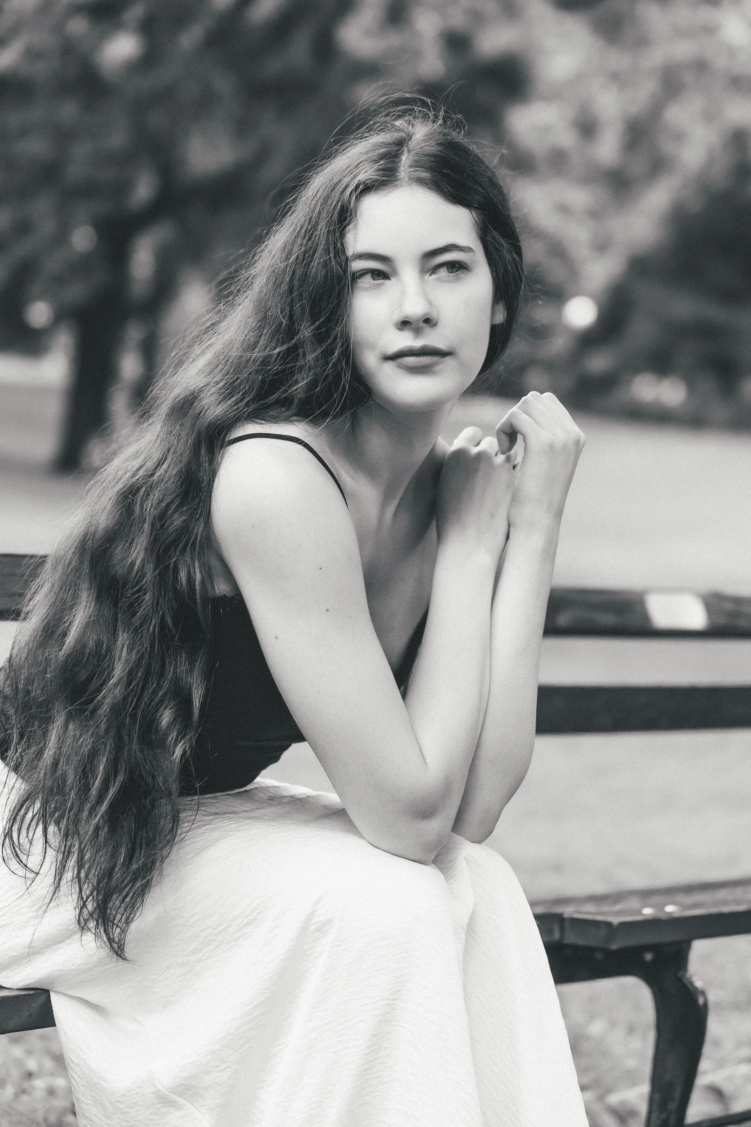 Black and white photo of a woman with long, wavy hair sitting on a bench outdoors, with a blurred background of trees.
