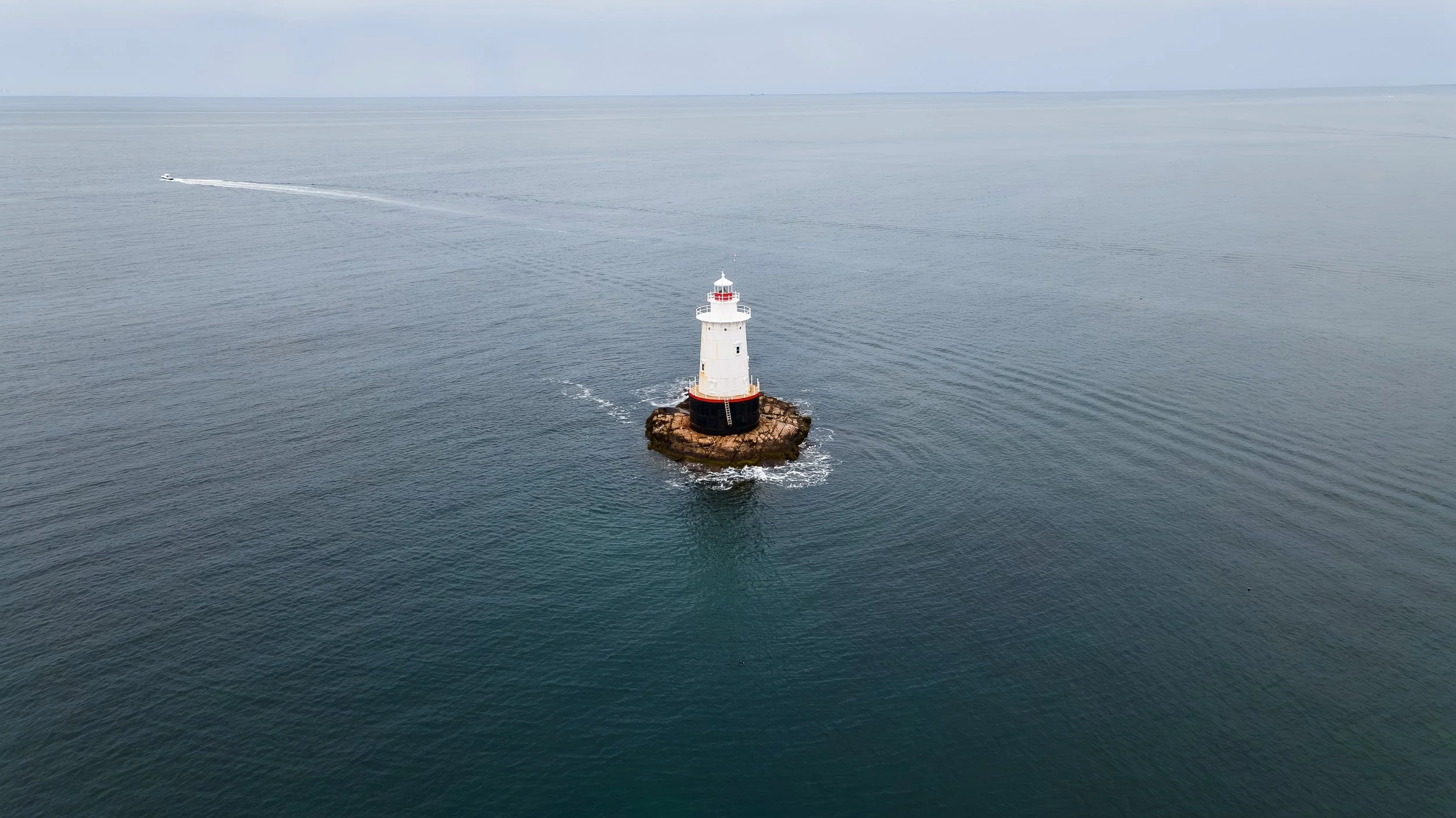 An aerial view of a white lighthouse on a rocky island, surrounded by calm ocean waters, with a boat passing nearby leaving a wake behind.