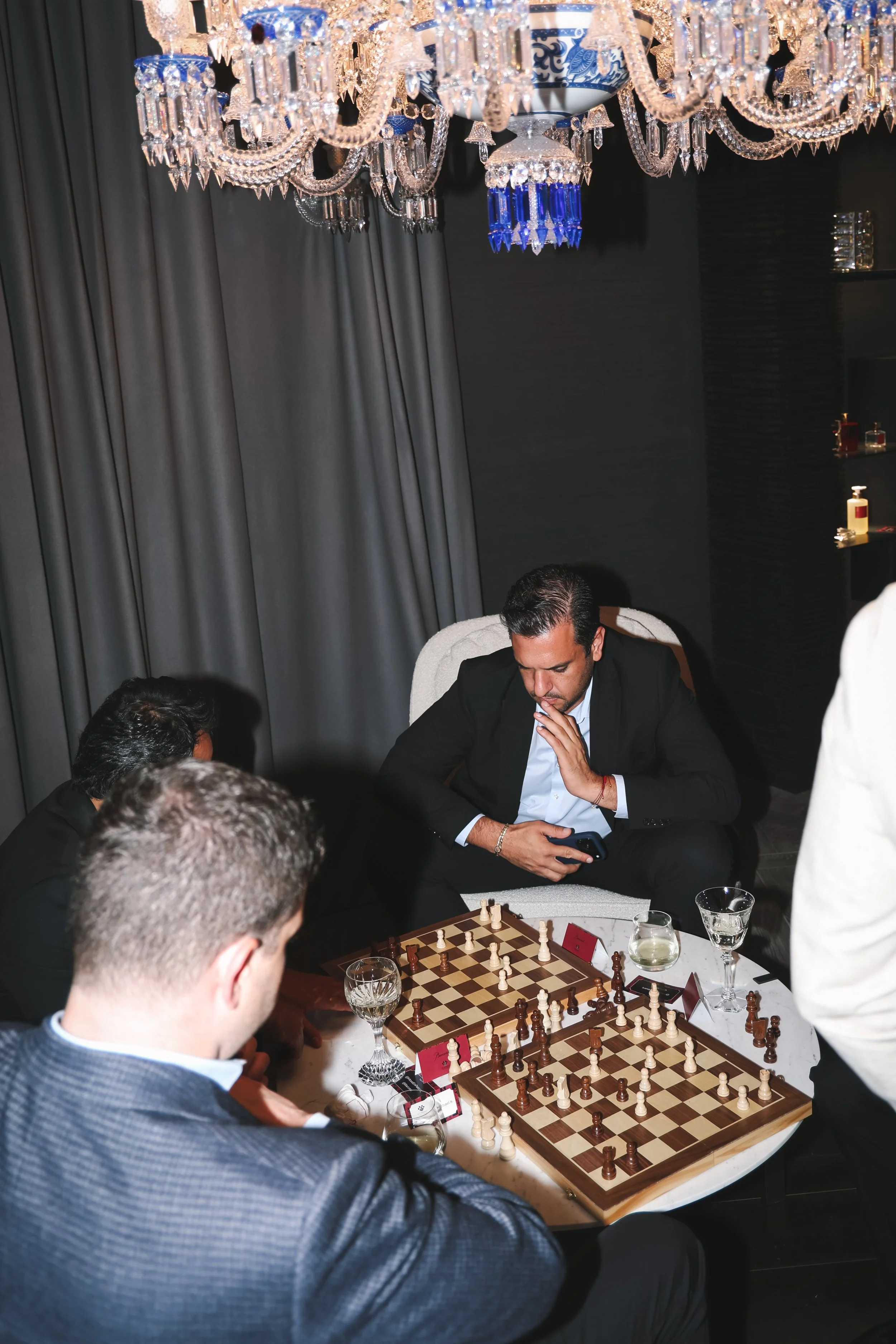 Men playing chess at a formal gathering, seated around a chessboard on a white table, with glasses of drinks, a chess clock, and a decorative chandelier above. The room has dark curtains and shelves with items.