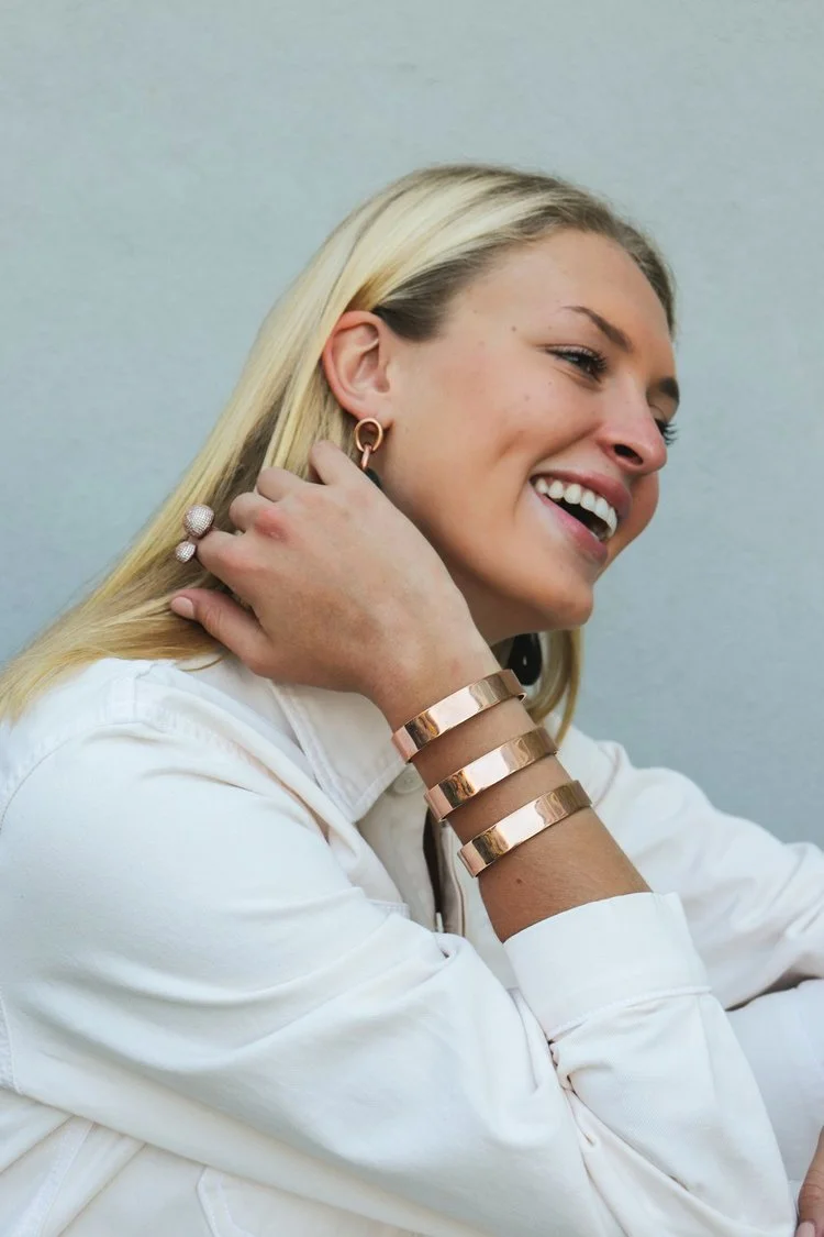 A woman with blonde hair wearing a white shirt, gold and leather bracelets, and earrings, smiling and touching her neck against a light background.