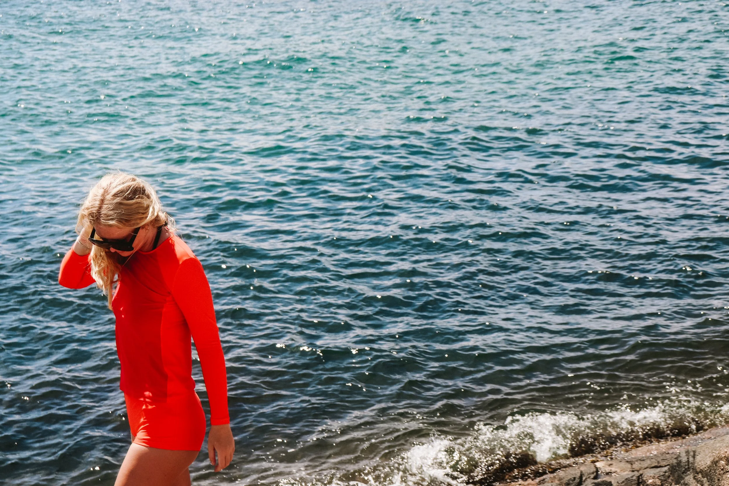 Woman in a red dress and sunglasses walking along a rocky shoreline with water in the background.