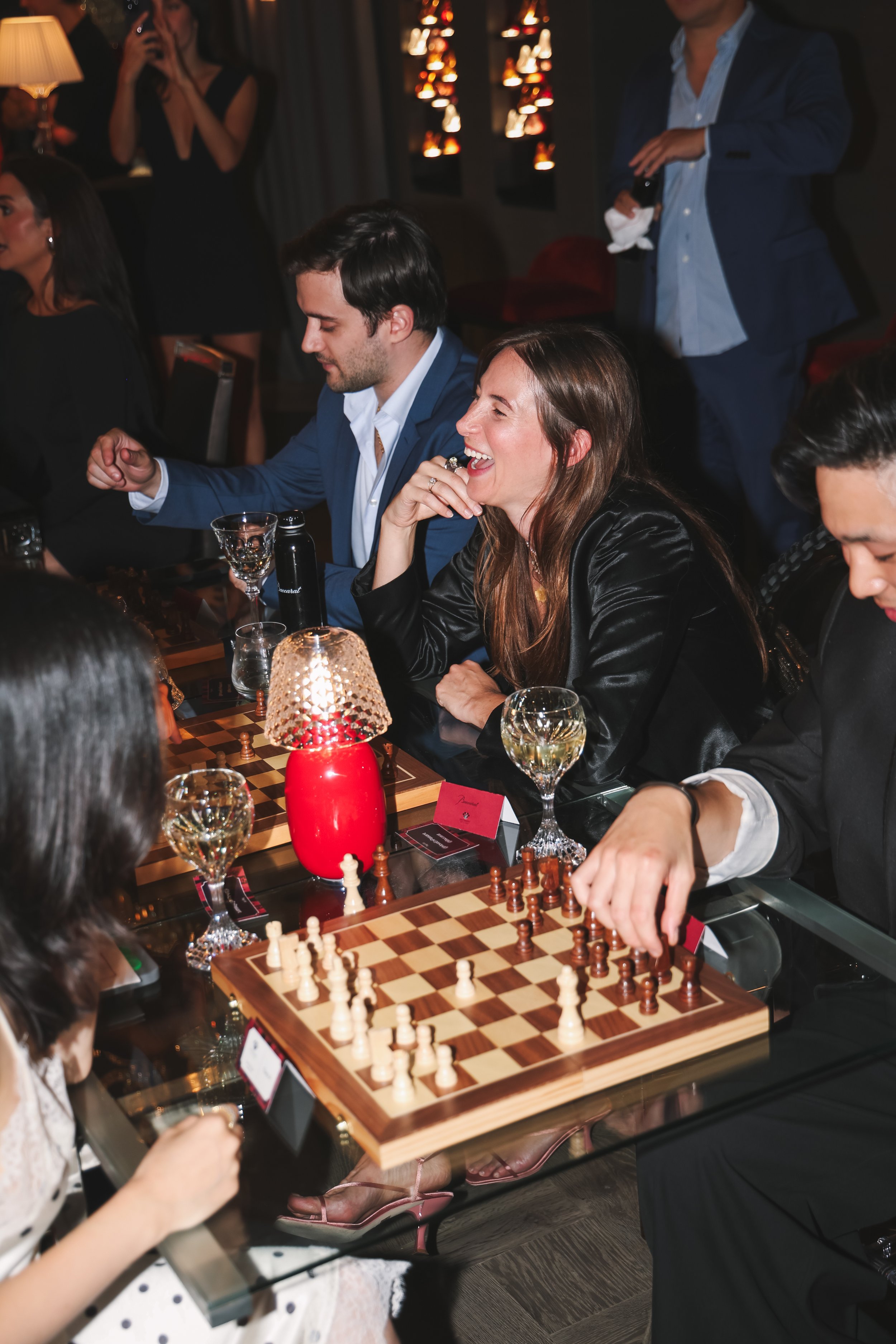People playing chess at a social gathering, with others watching and taking photos, in a cozy, dimly lit room.