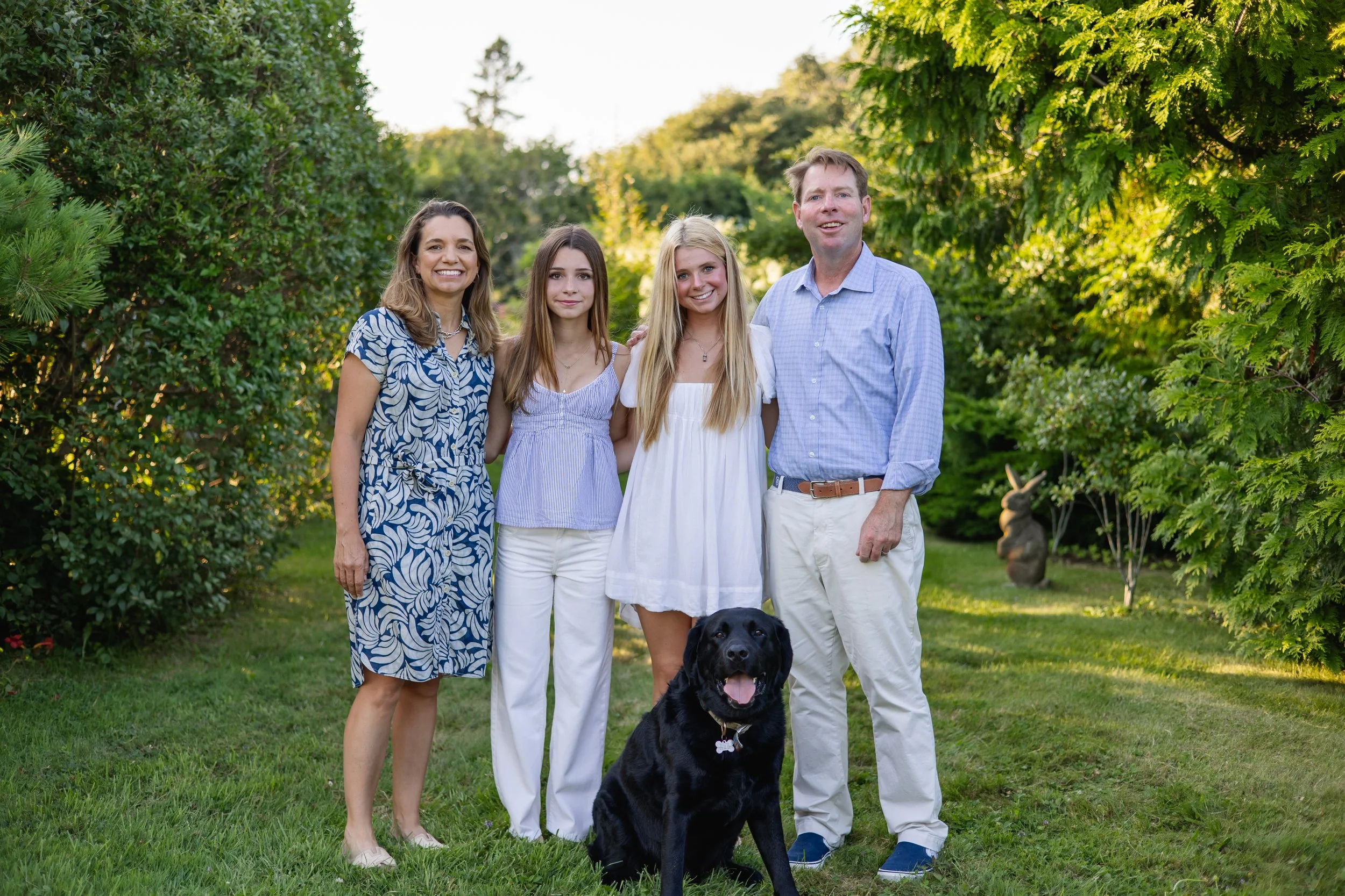 A family of five, including three young women, one man, and a black dog, standing together outdoors among green trees and bushes, smiling at the camera.