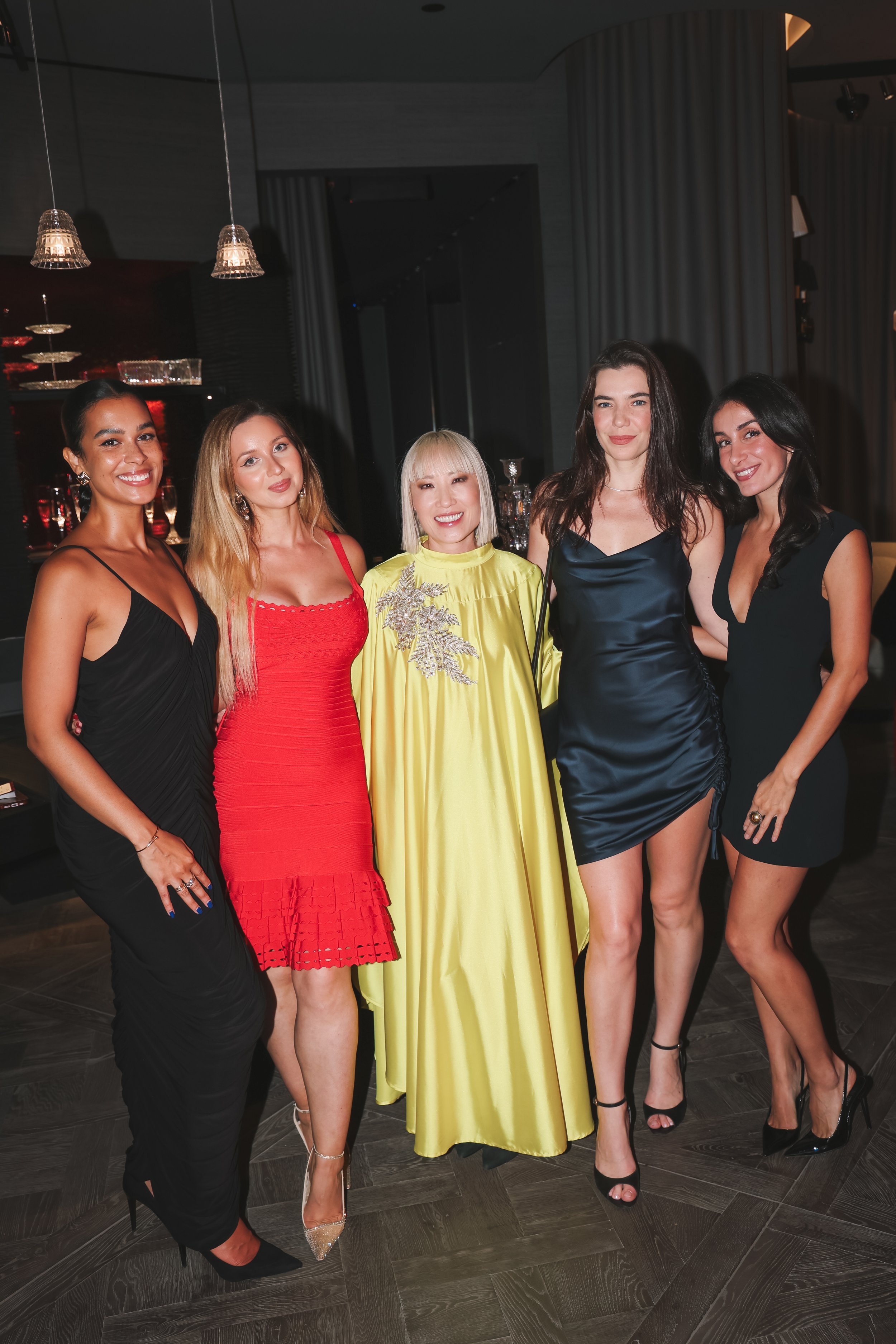Group of five women dressed in formal evening wear, standing together in a modern, dimly lit venue.