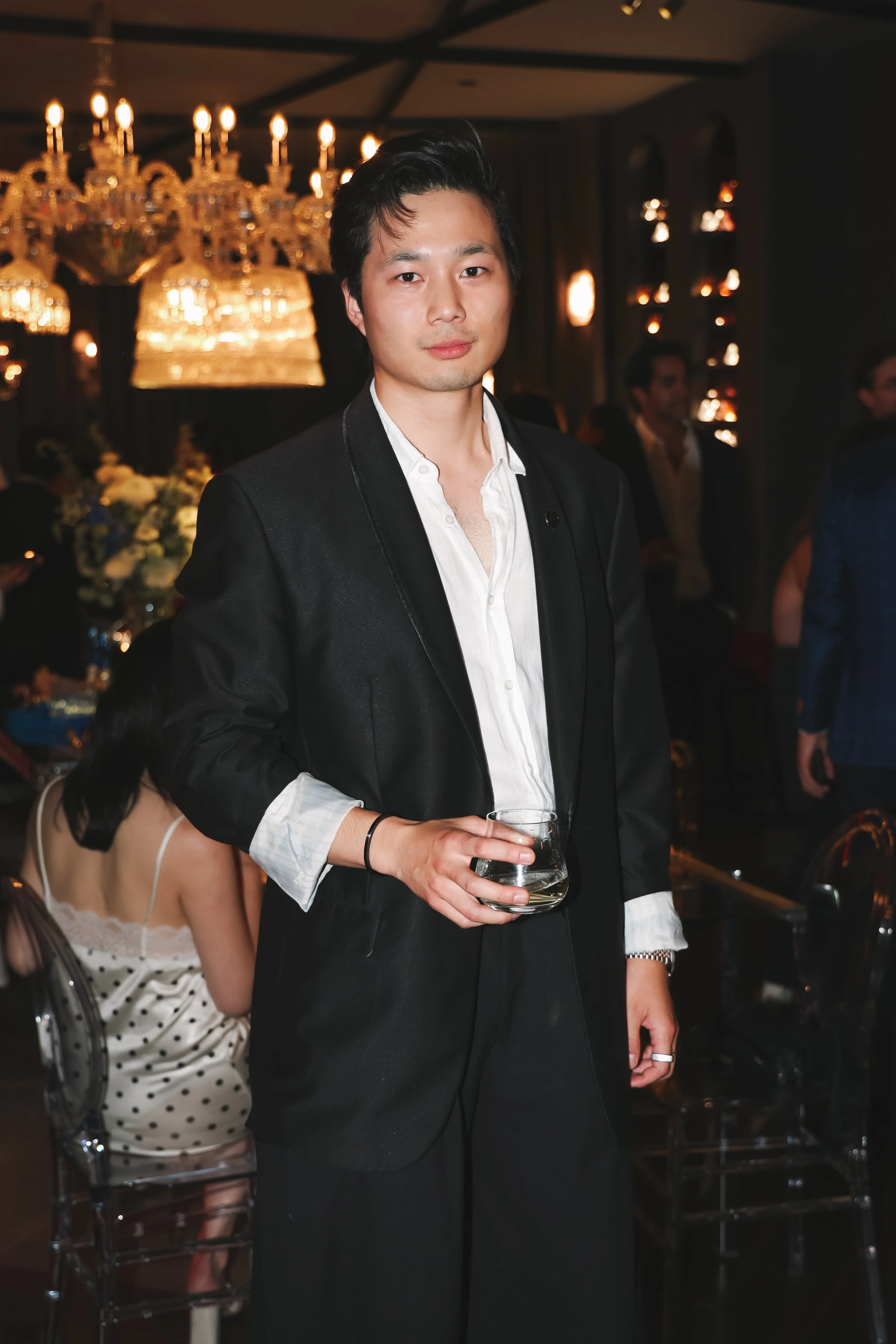 A young man in a black suit holding a glass of wine at a fancy indoor party or event with warm lighting, chandeliers, and other people in the background.