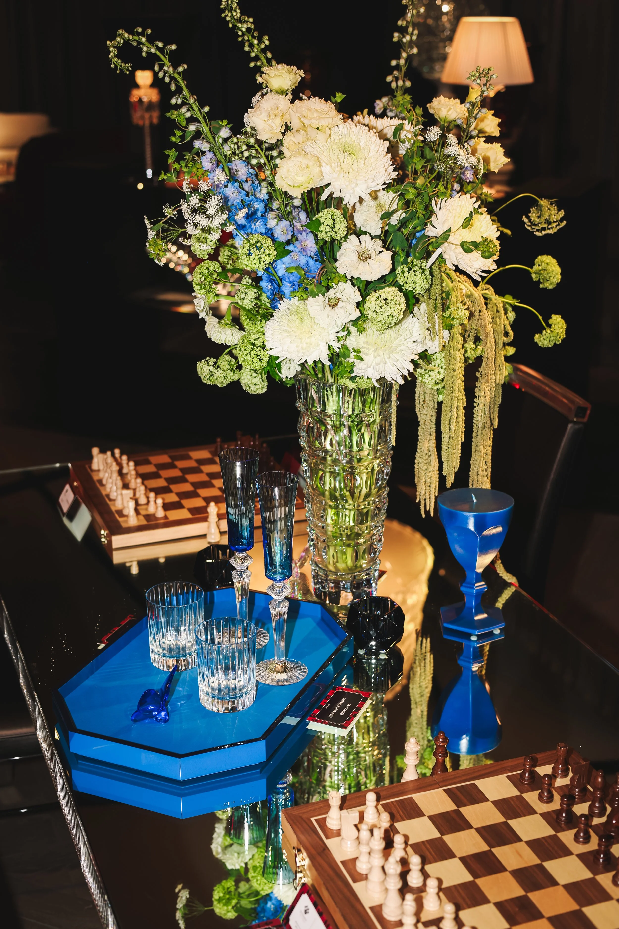 A tall clear glass vase holds a large bouquet of white, cream, blue, and green flowers, including chrysanthemums, hydrangeas, and various greenery, arranged on a shiny black table in a dimly lit room with a beige lamp in the background.