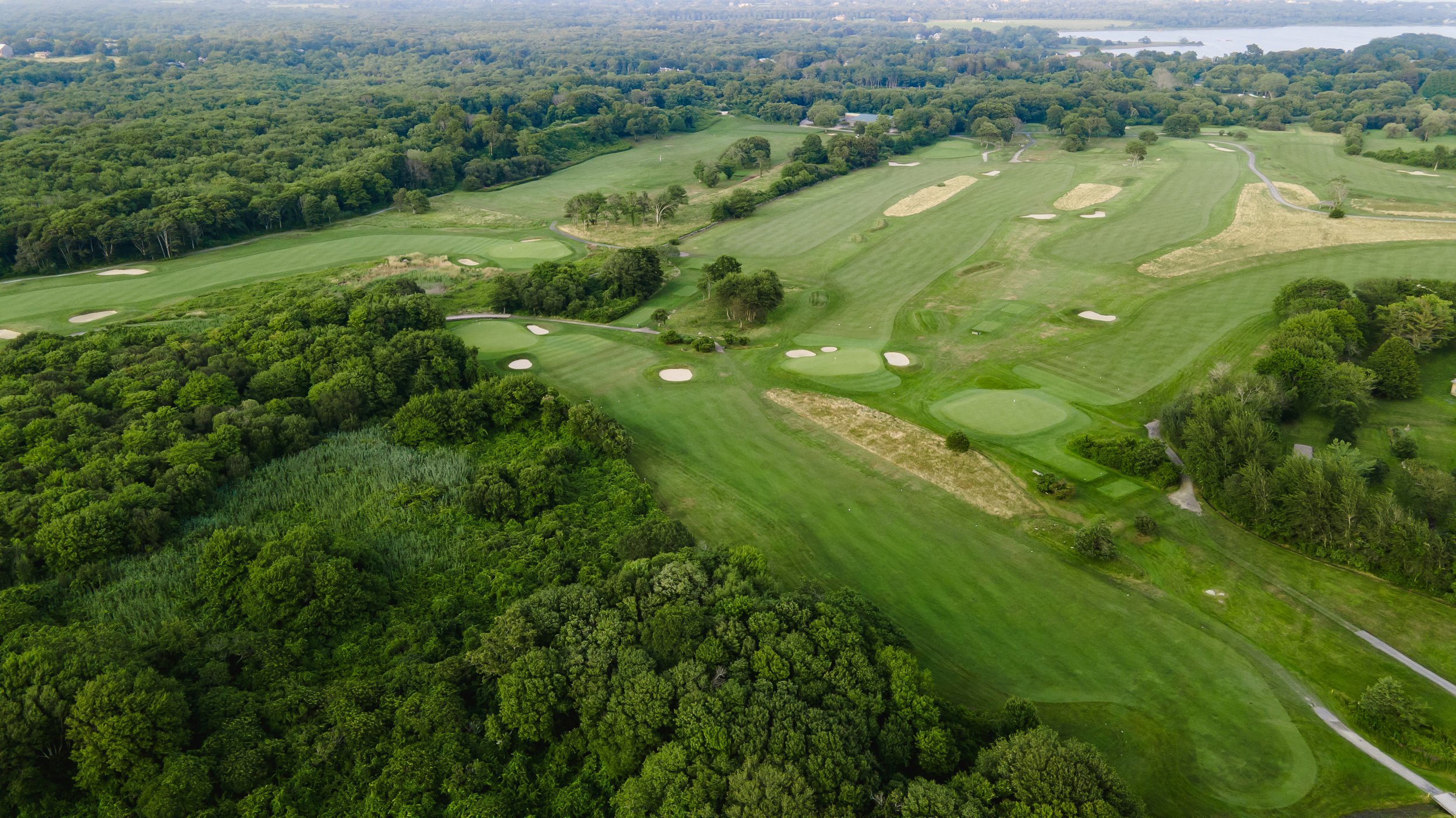 Aerial view of a golf course surrounded by lush green trees, with fairways, sand traps, and greens visible, and a body of water in the distance.