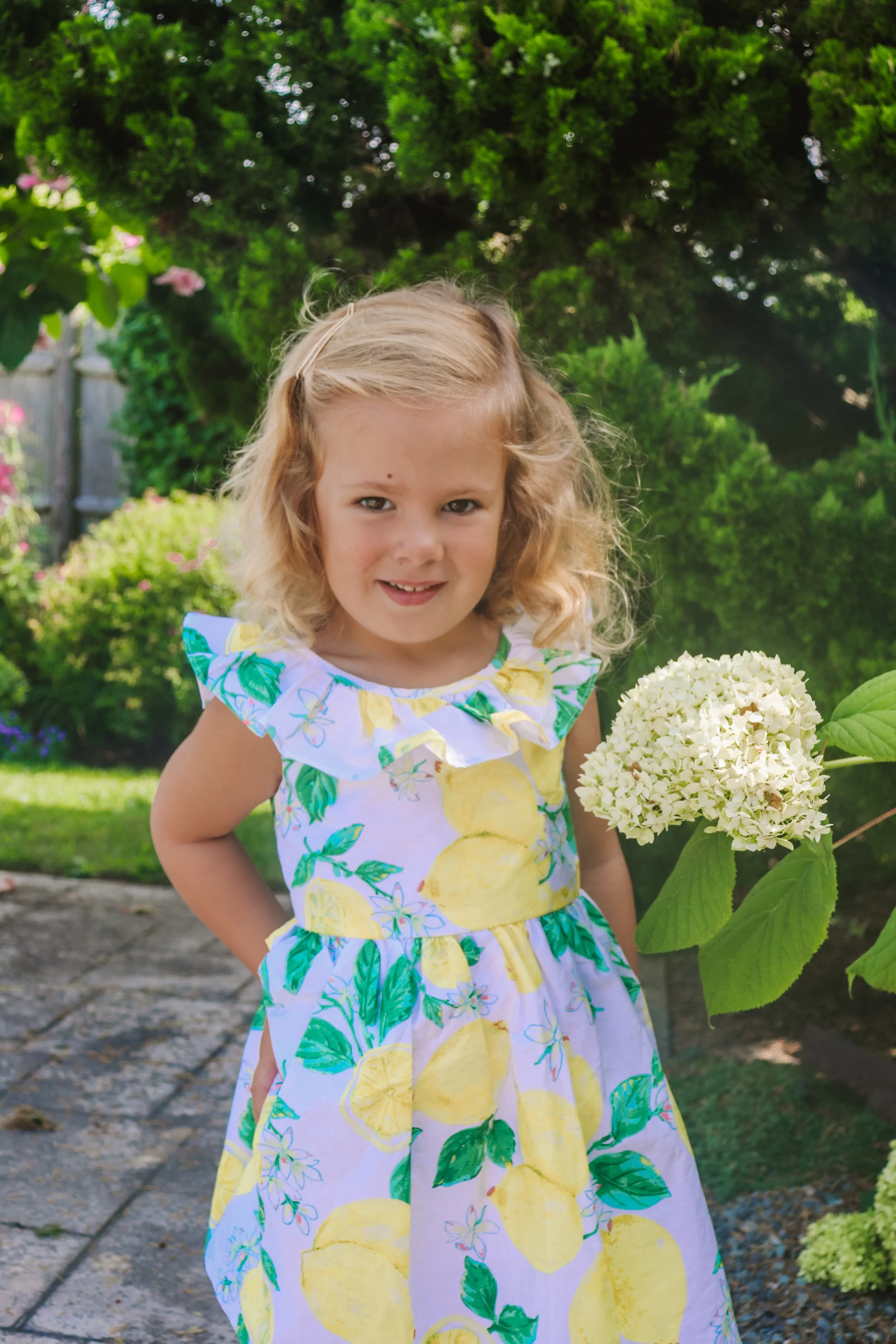 A young girl with blonde curly hair wearing a dress with lemon and lemon leaf pattern, standing outdoors in a garden with green trees and bushes, holding a white hydrangea flower.