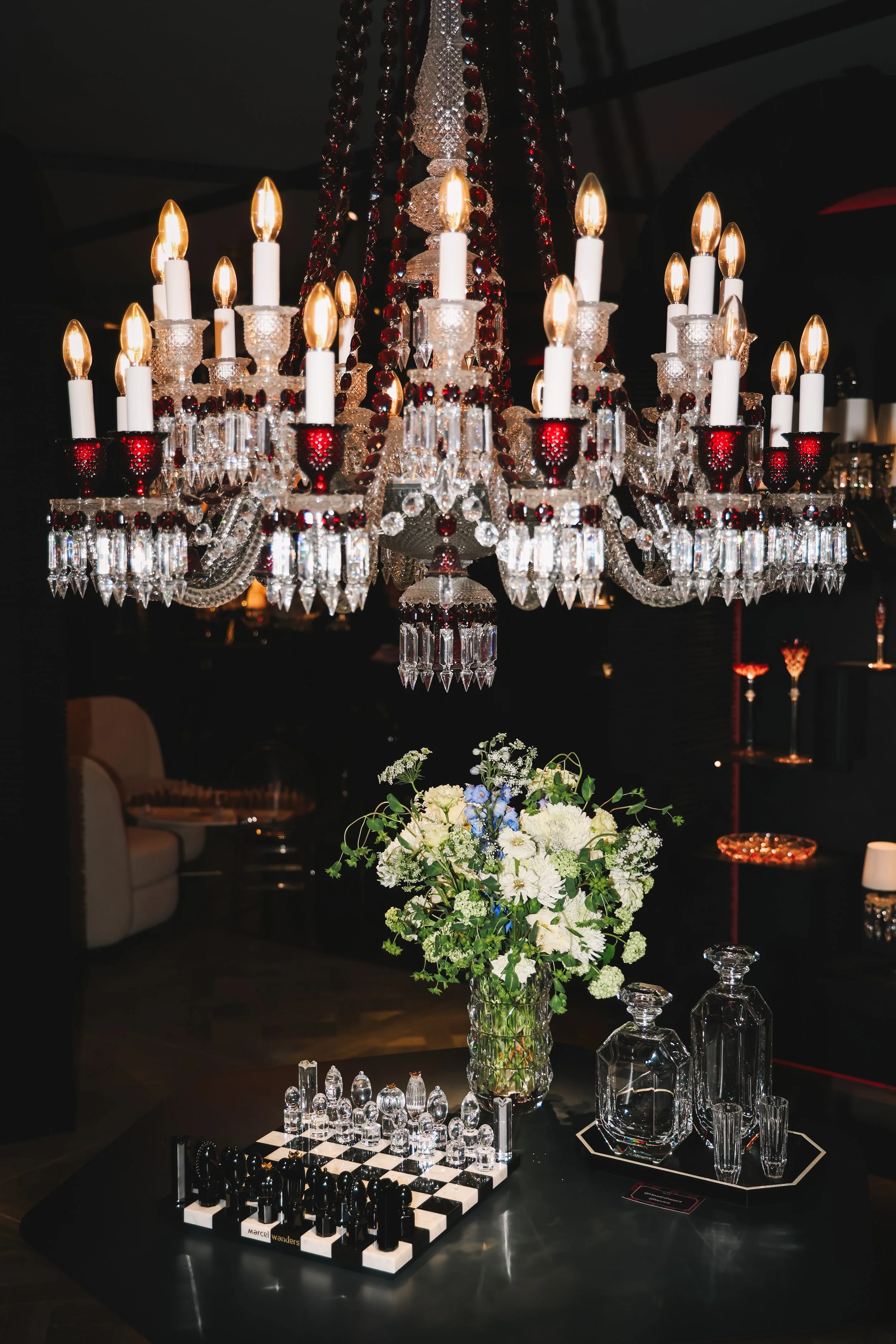 A grand chandelier with candle-shaped lights, crystal ornaments, and red accents hangs above a table. Below, a glass vase with a white and green flower arrangement, and a chessboard set with chess pieces are displayed on a dark table.