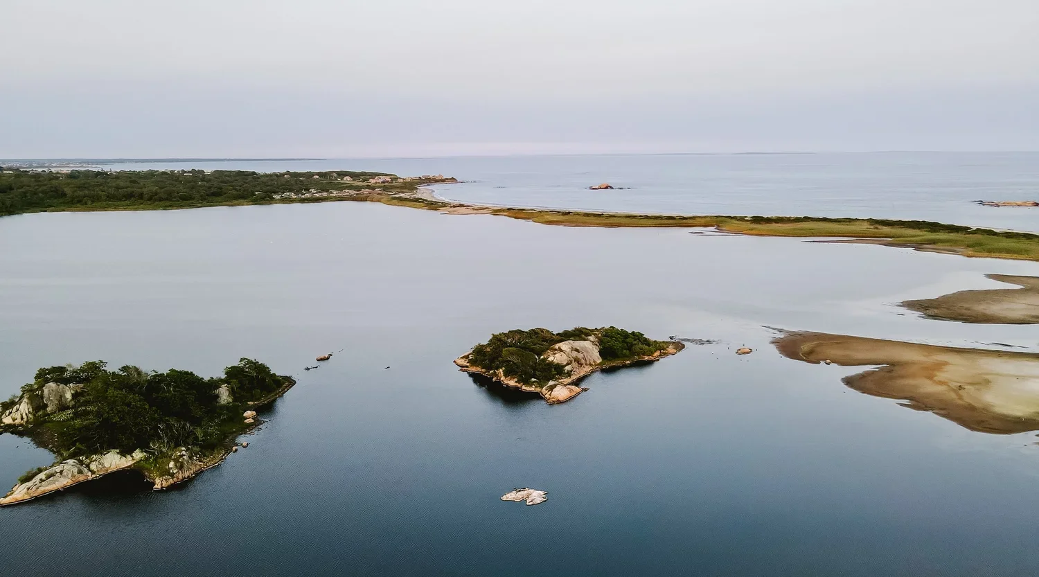 Aerial view of a coastal landscape with small islands, calm water, sandbars, and lush green vegetation, with a shoreline and residential area in the distance under an overcast sky.