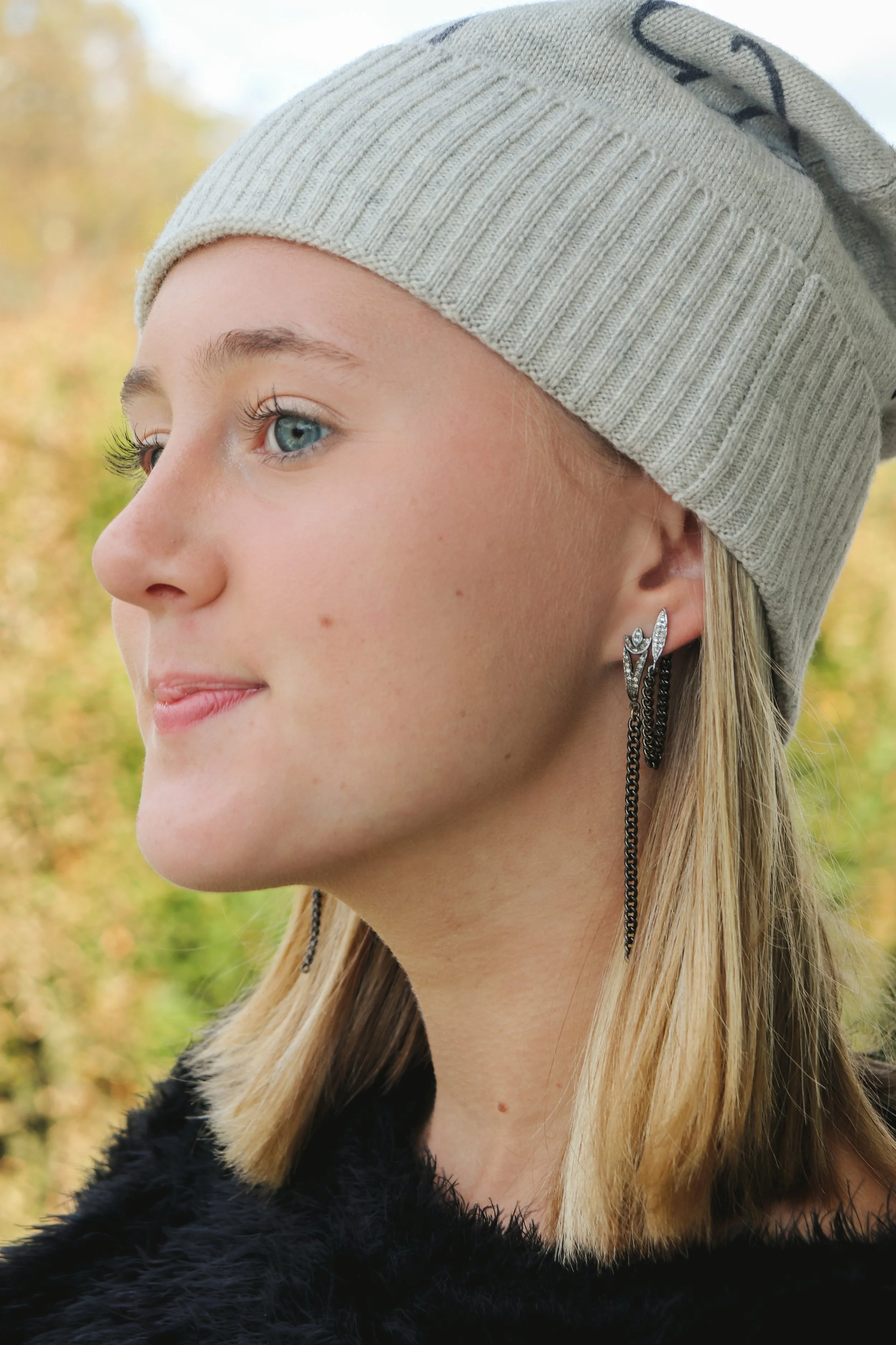Close-up of a young woman with blonde hair, wearing a gray knit beanie and black fuzzy coat, looking to the side against a background of blurred autumn trees.
