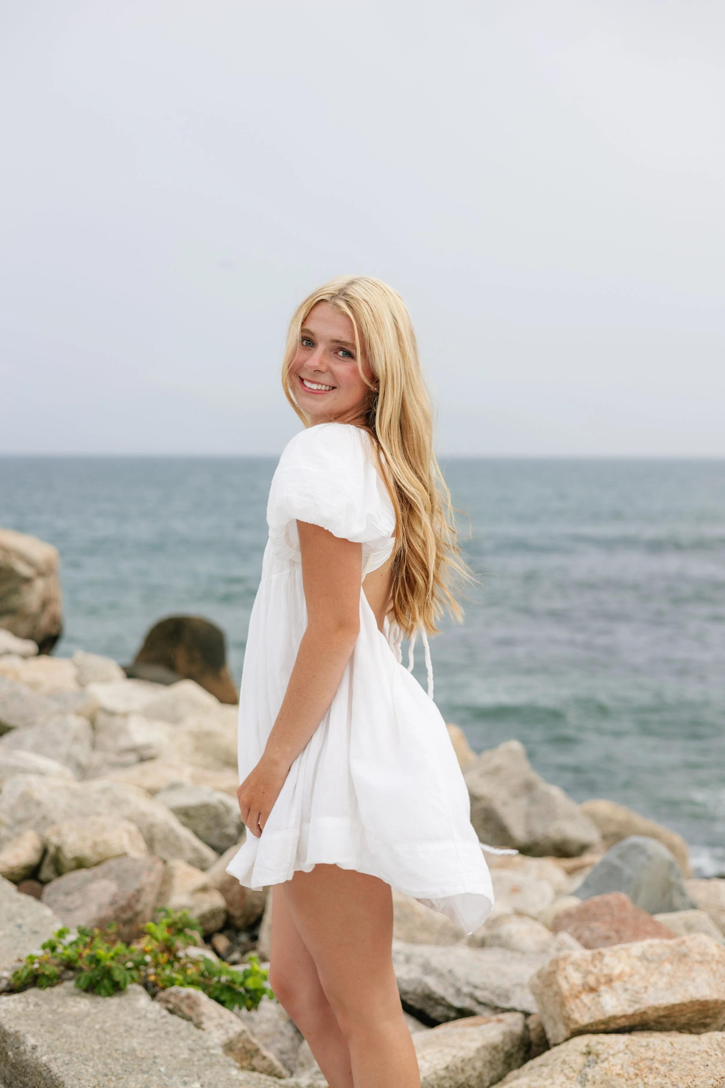 A young woman wearing a white summer dress with short sleeves, standing on rocks by the ocean, smiling at the camera with long blonde hair, cloudy sky background.