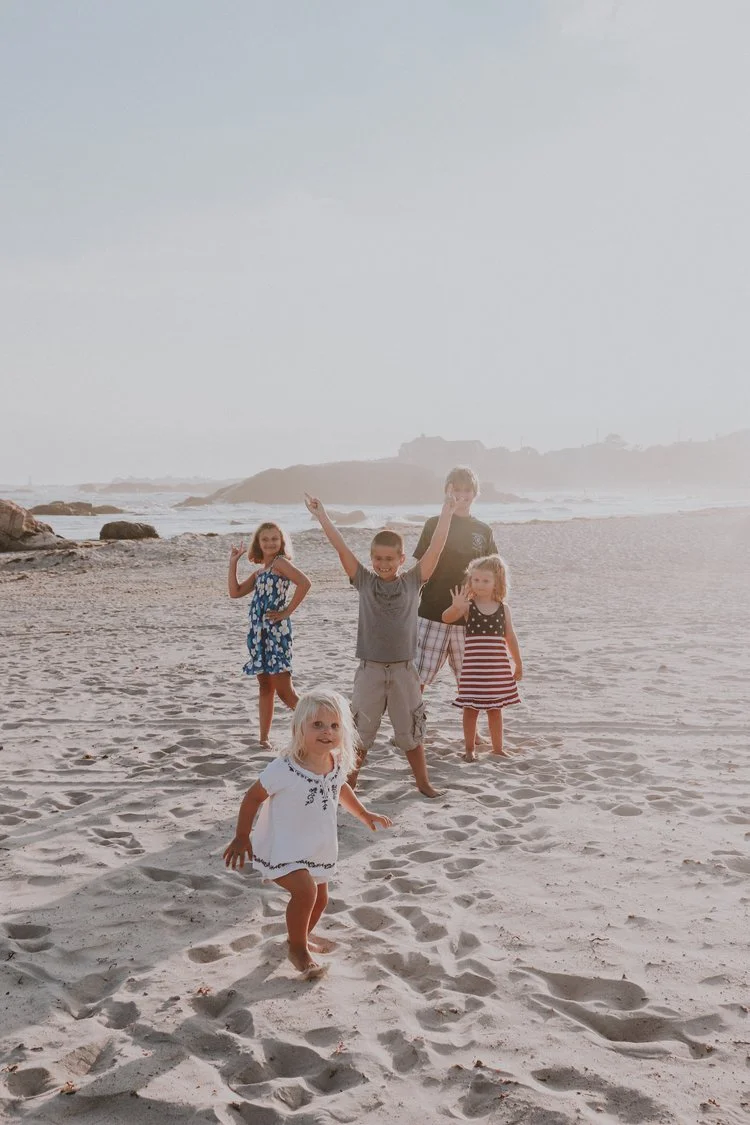 Group of five children, four girls and one boy, on a sandy beach during daytime. The children are standing and posing, with some making gestures, and the ocean and rocks are visible in the background.