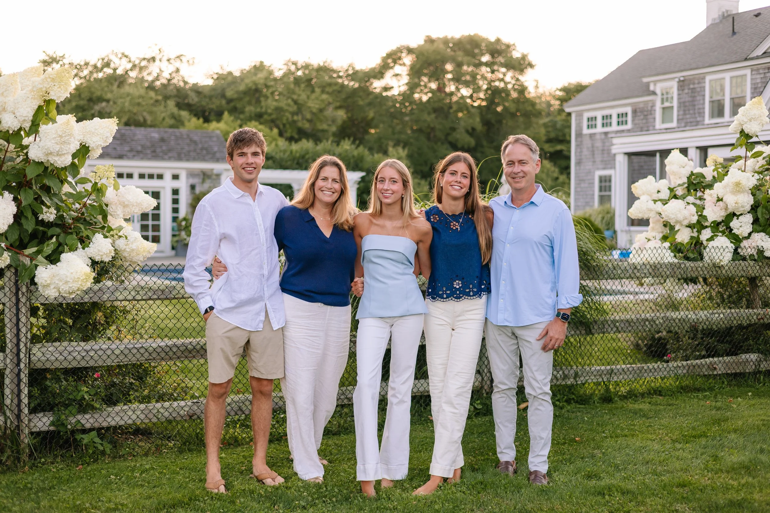 A family of five standing together outdoors in front of a black fence, with green trees, shrubs, and flowering plants around them, and a large house behind them. The family is dressed in semi-formal attire and smiling at the camera.