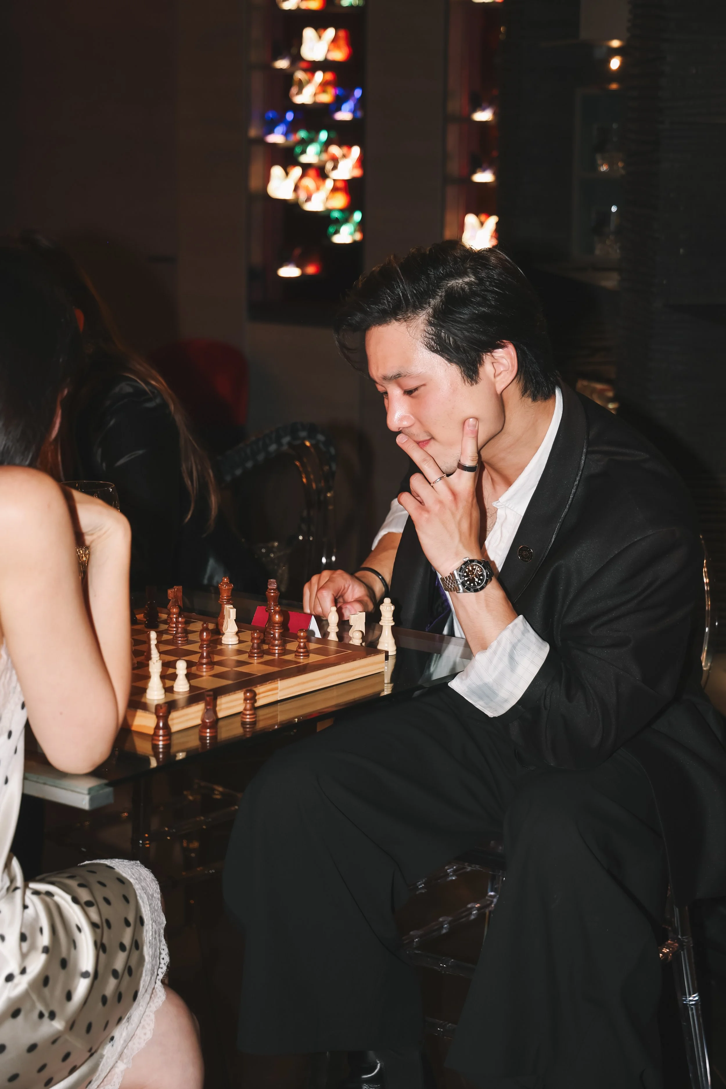 A young man wearing a black suit, white shirt, and black watch is playing chess at a lounge or bar. He is touching his chin and looking at the chessboard, with a thoughtful expression. The background is blurred, showing colorful lights and decorated 