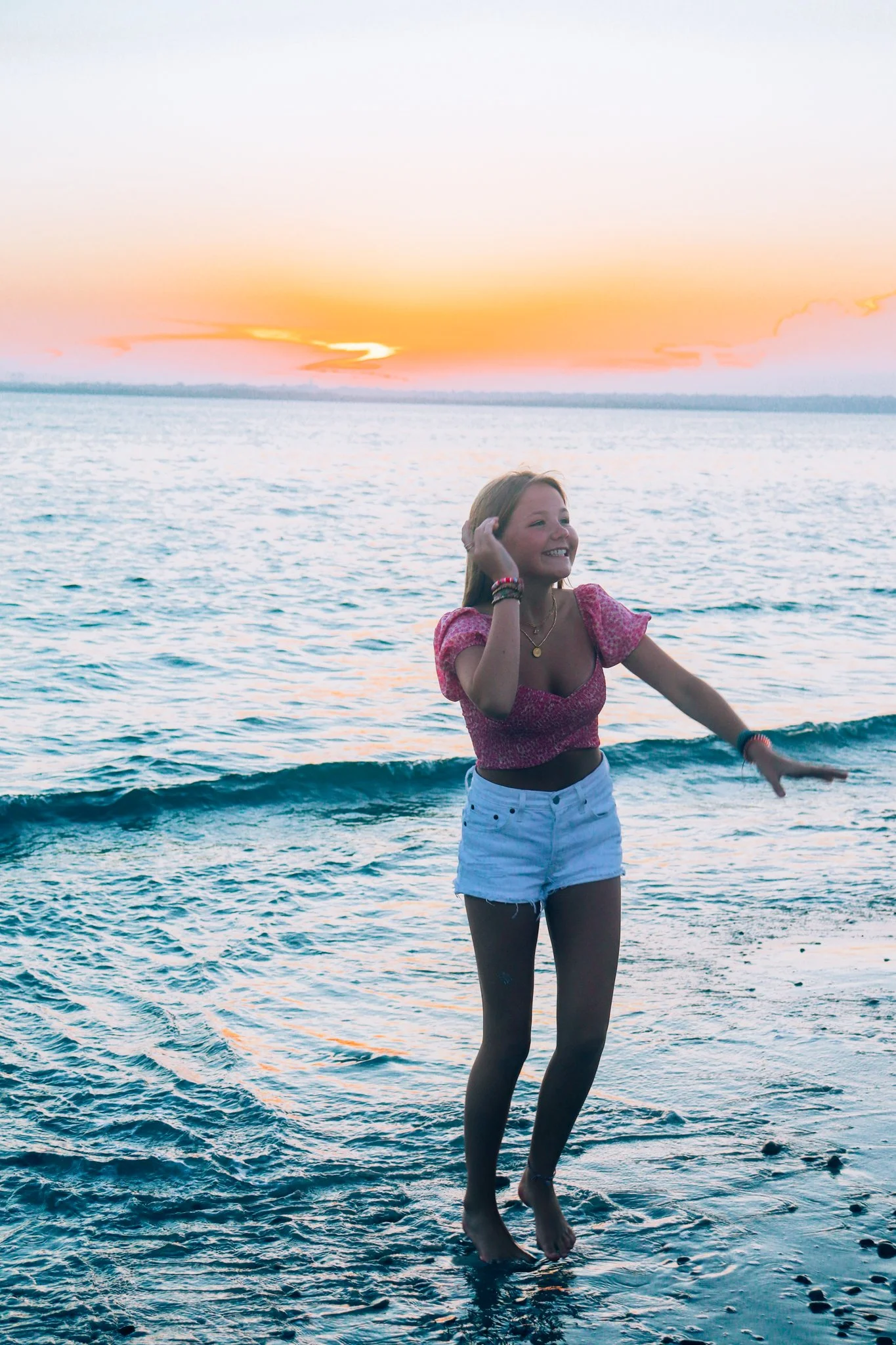 A young woman with long hair, wearing a pink crop top and white shorts, standing in shallow ocean water at sunset, smiling and playing.